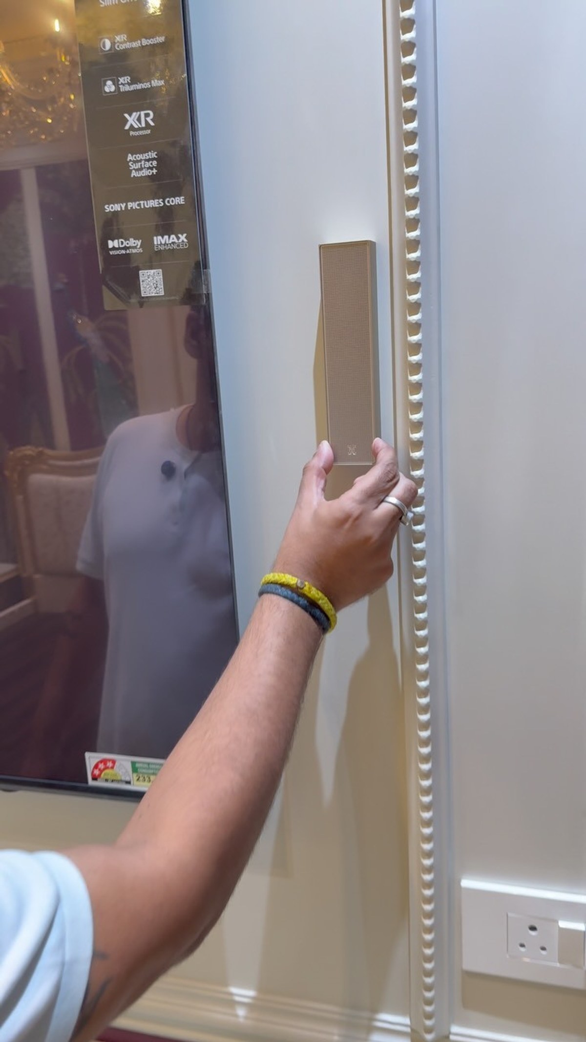 Karan Sethi of Qube Designs touching a champagne XSCACE Cane slim array speaker mounted on a cream moulded wall panel beside a large television