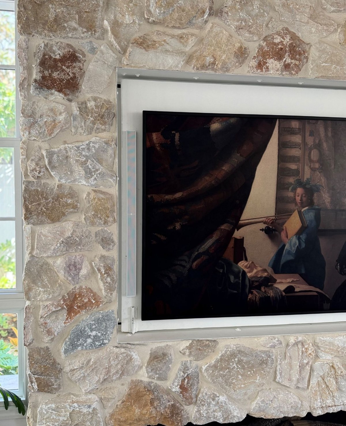 Daylight from a window falls across the limestone wall, with a slim white QuadCane speaker column visible against the white reveal beside the recessed television