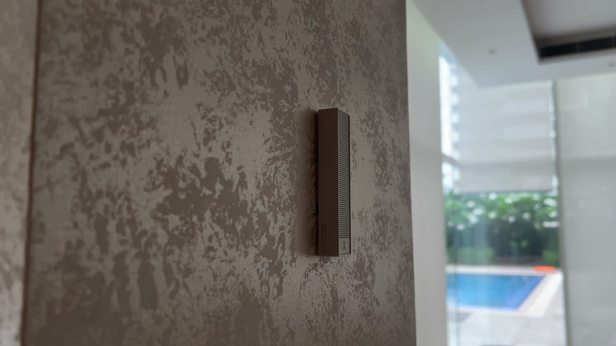 A white XSCACE Bonsai seen at a raking angle showing its 12mm depth and side branding, on a textured plaster pier with a swimming pool visible through the glazing beyond