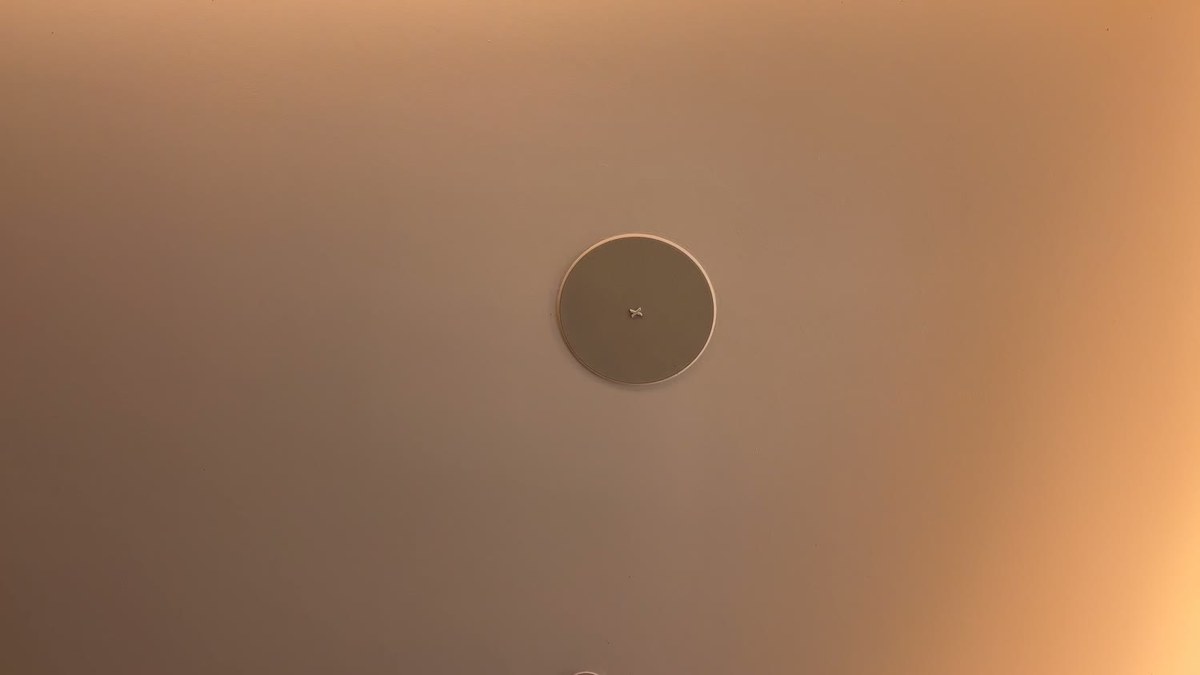 Close-up of a flush-mounted XSCACE Aspen in-ceiling speaker painted to match the ceiling, with the XSCACE mark at its centre, lit by warm cove lighting
