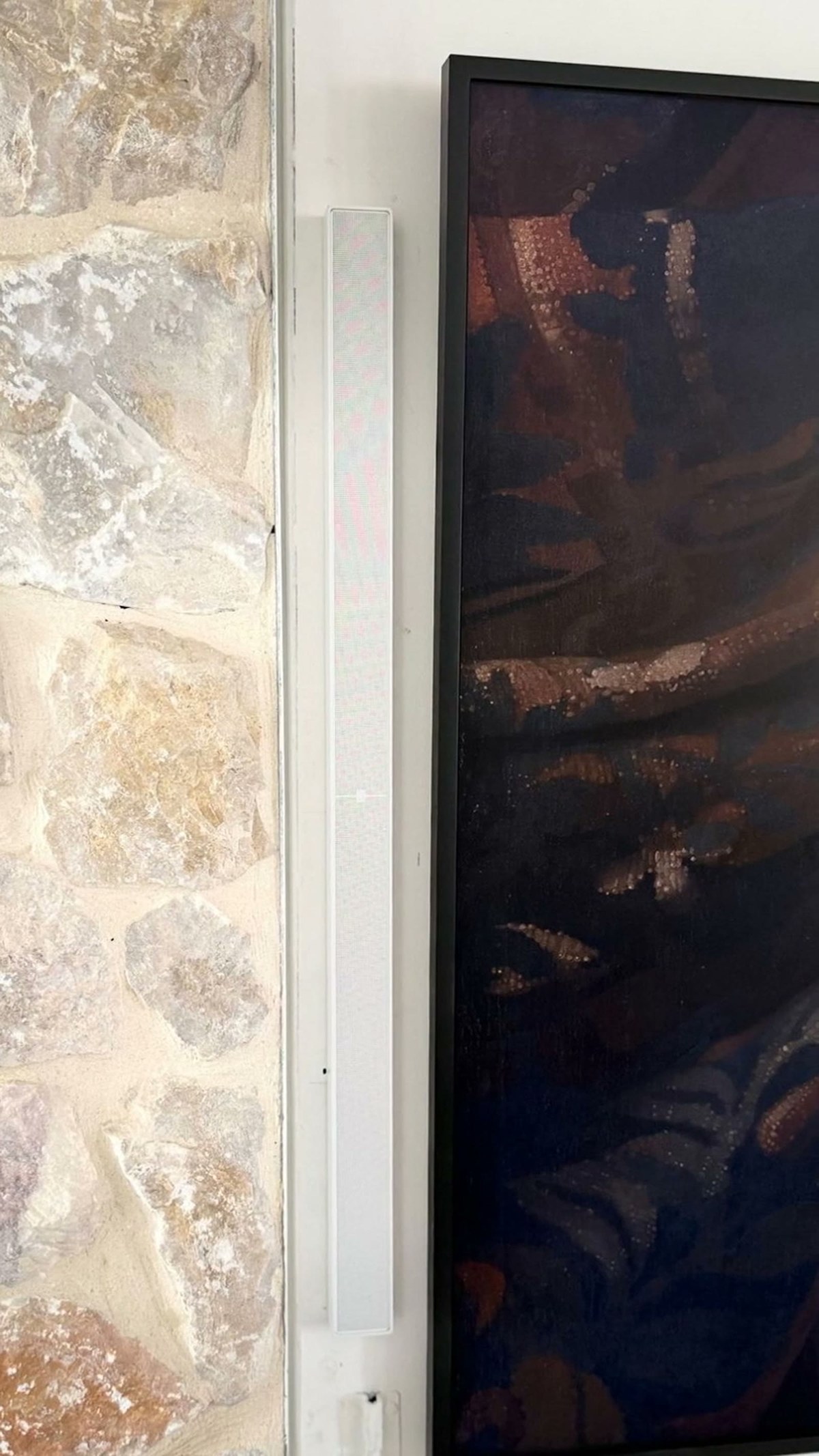 A close view of a white XSCACE QuadCane slim array speaker mounted beside a recessed television, pulling back to reveal the limestone fireplace wall