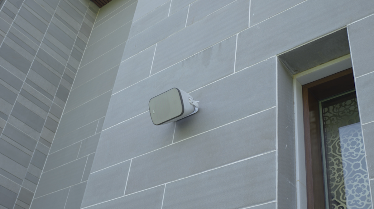 An XSCACE Camphor 6 all-weather outdoor patio speaker securely bracket-mounted horizontally to a wooden architectural pillar in a modern industrial-style outdoor seating environment.