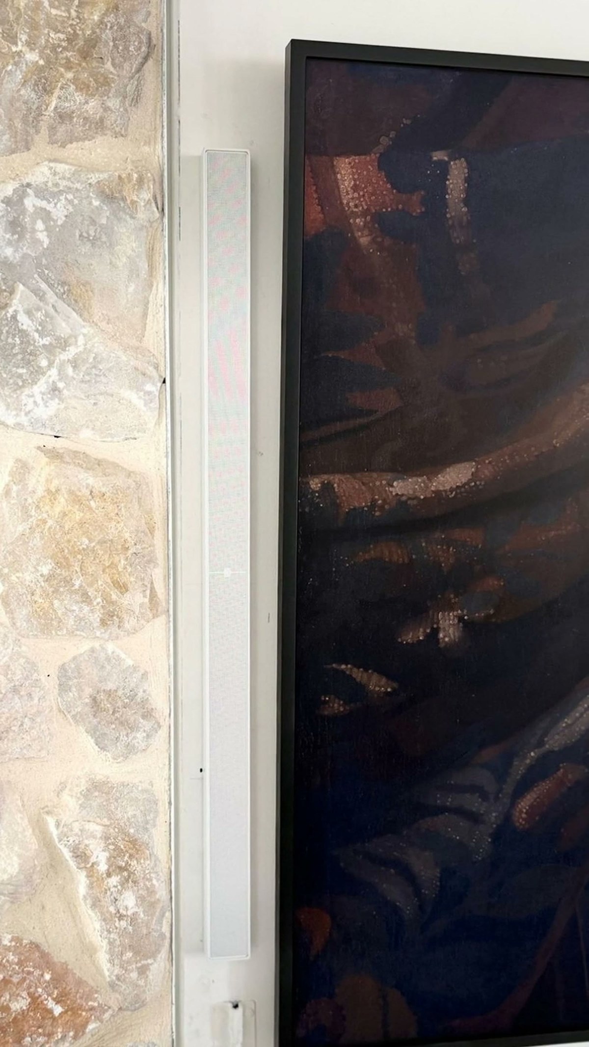 A close view of a white XSCACE QuadCane slim array speaker mounted beside a recessed television, pulling back to reveal the limestone fireplace wall