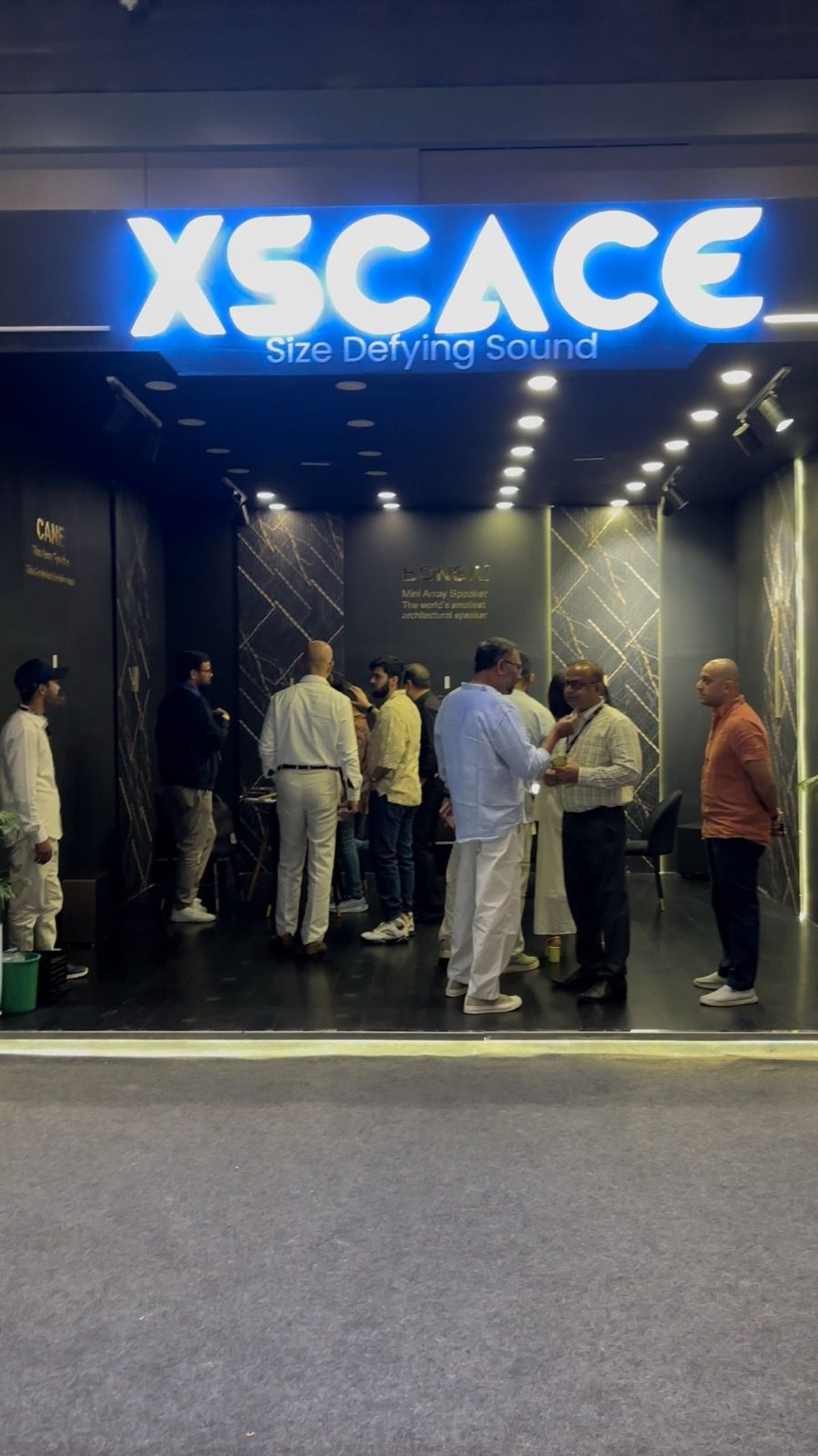 The entrance to the XSCACE stand at the Architectural Digest Design Show 2025, with an illuminated blue XSCACE Size Defying Sound sign above a dark walk-in space lined with marble slat walls