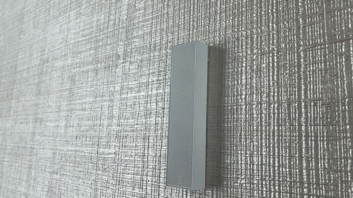 Close-up of a white XSCACE Cane slim array speaker on textured plaster, seen at a raking angle showing its 23mm depth and perforated grille