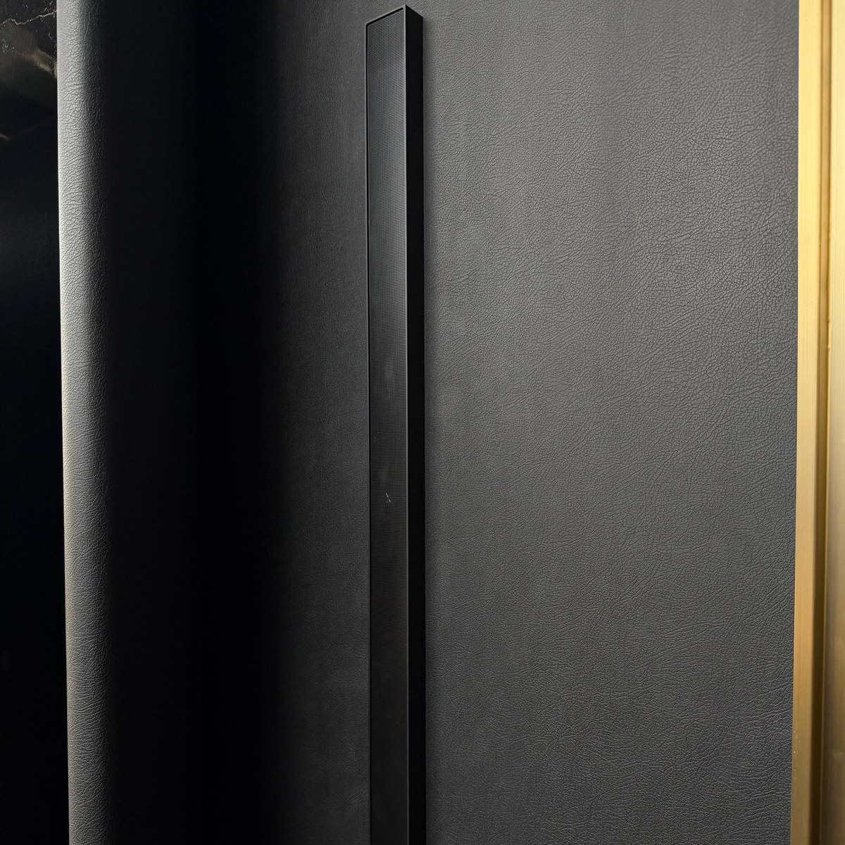 XSCACE QuadCane speaker mounted on black leather wall panelling beside a brass inlay strip