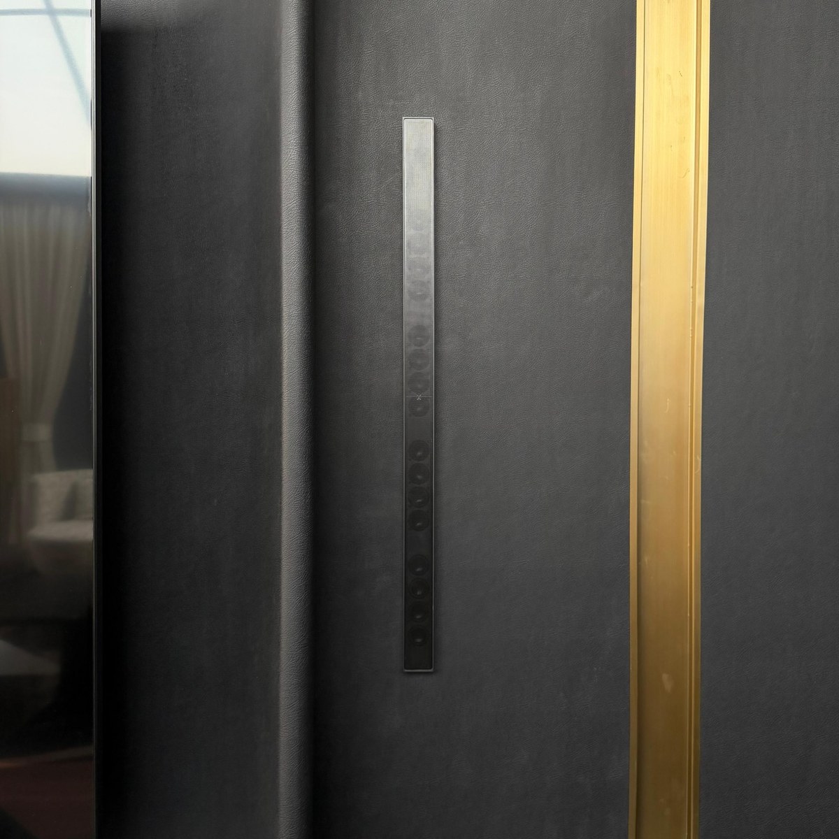 XSCACE QuadCane speaker mounted on black leather wall panelling beside a brass inlay strip
