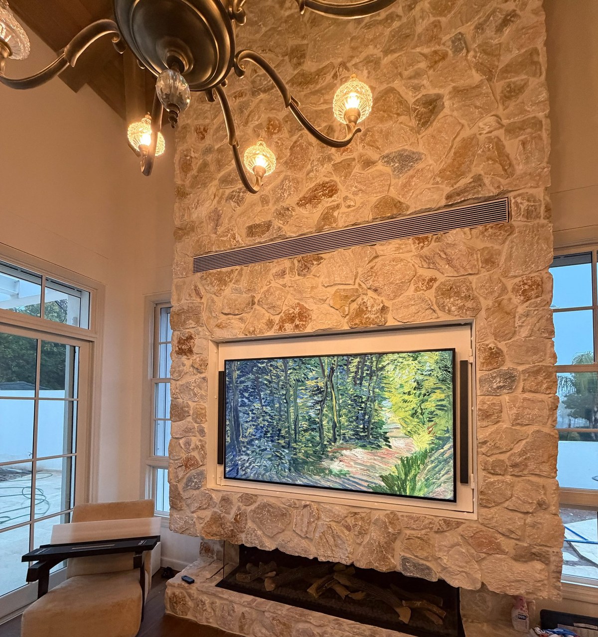 A double-height living room in Israel with a rough limestone chimney breast. A recessed art-mode display shows an oil painting of a woodland path, with a slim black XSCACE QuadCane speaker mounted vertically on the reveal at each side.