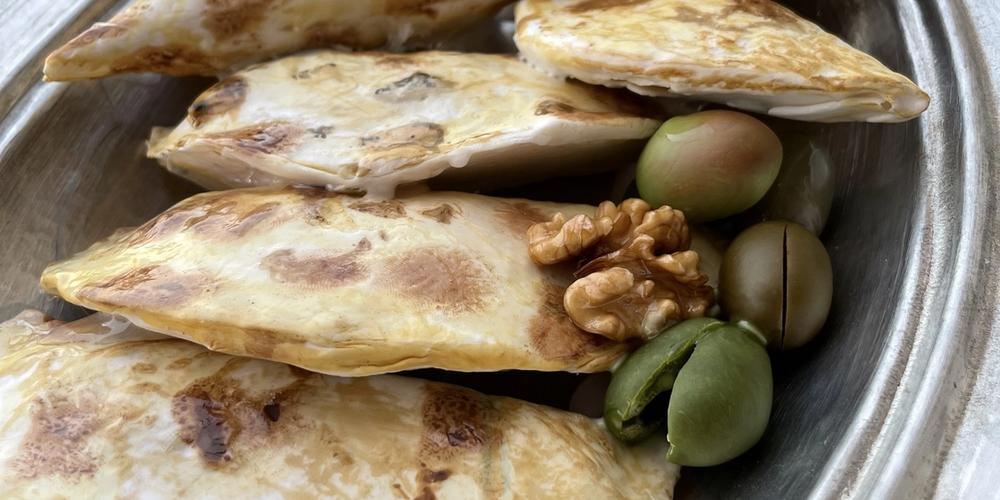 Oval metal tabakta üçgen zeytinli börek replikaları; kesilmiş parçalarda iç harç, yanında zeytin ve ceviz görülüyor.