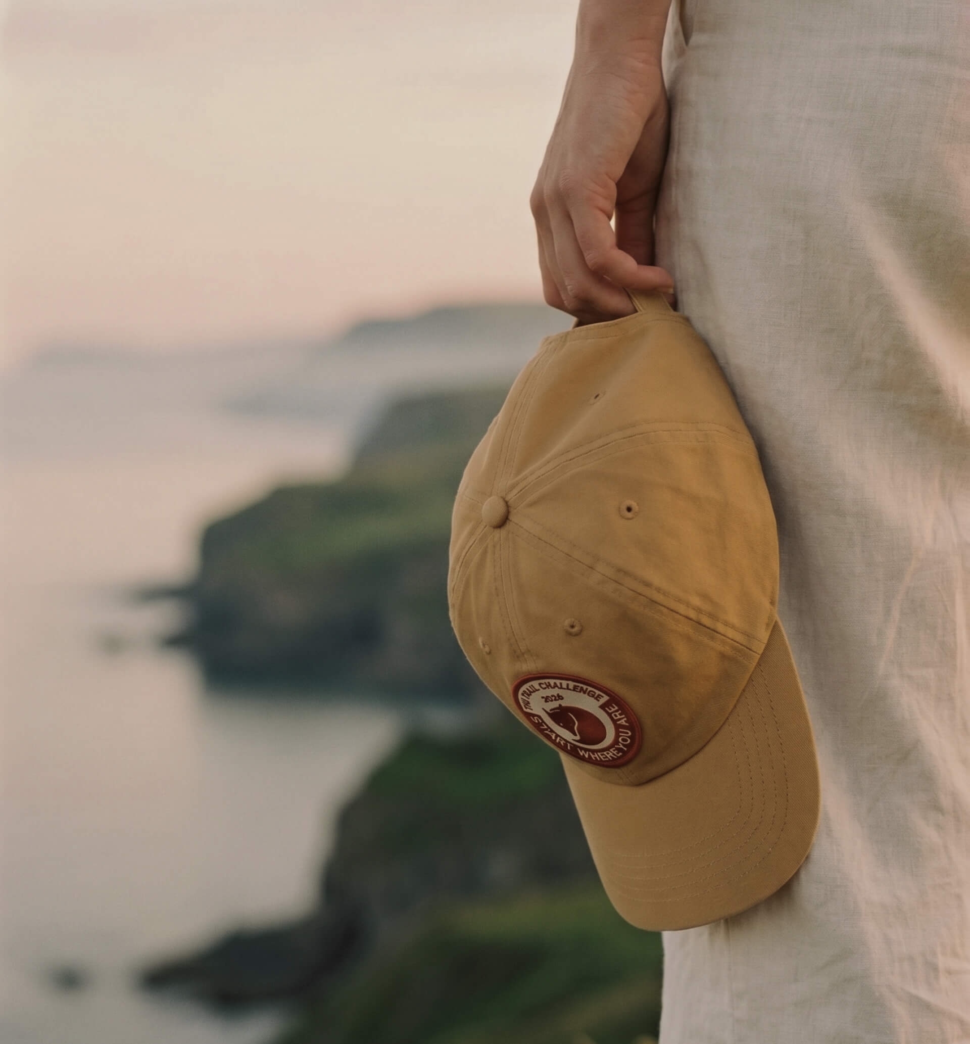 Side profile of a person holding a warm sand-colored cap with the Trail Challenge patch visible on the side panel.