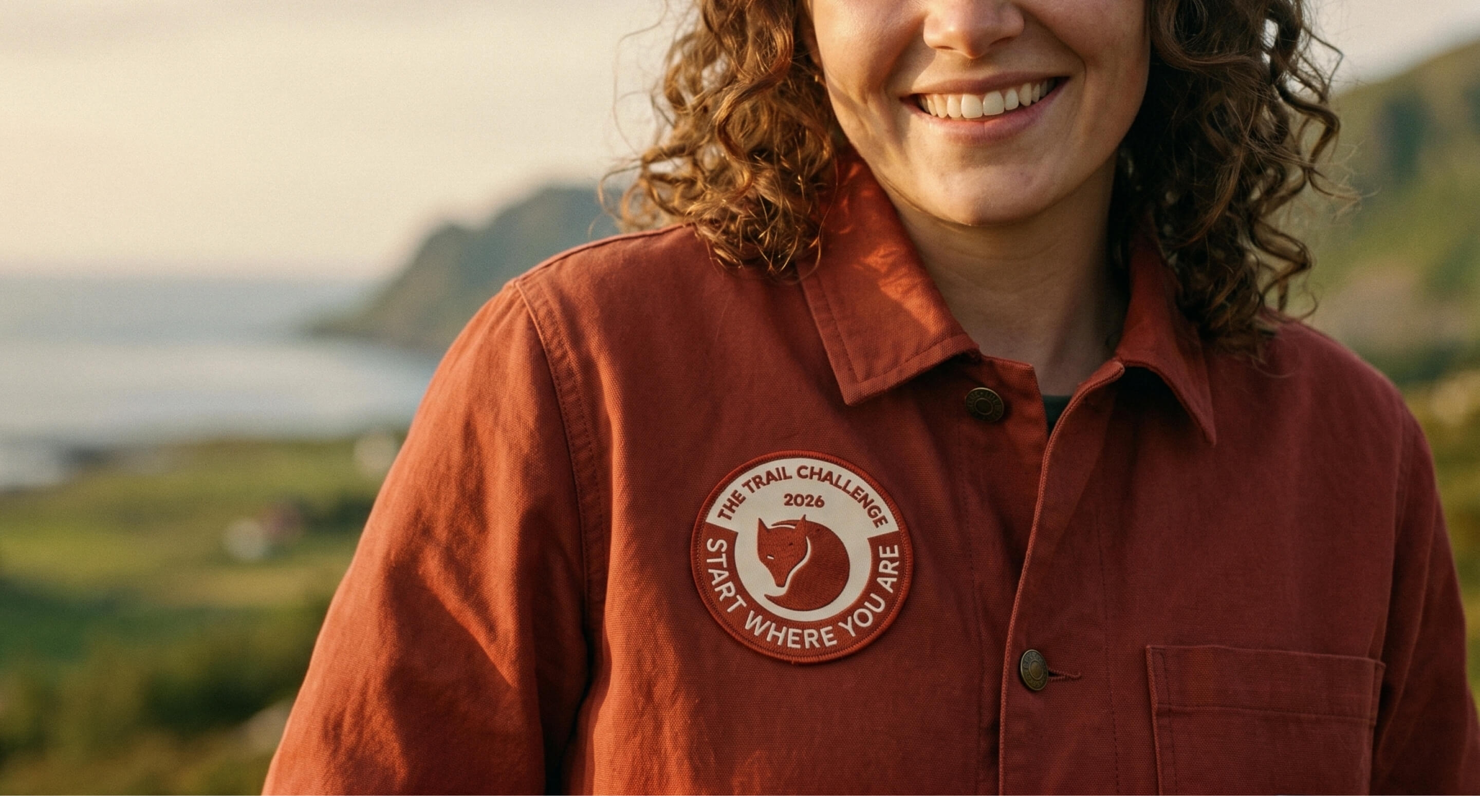 Close-up portrait of a woman with curly chestnut hair smiling, wearing a brick-red workshirt with the patch sewn onto the chest.