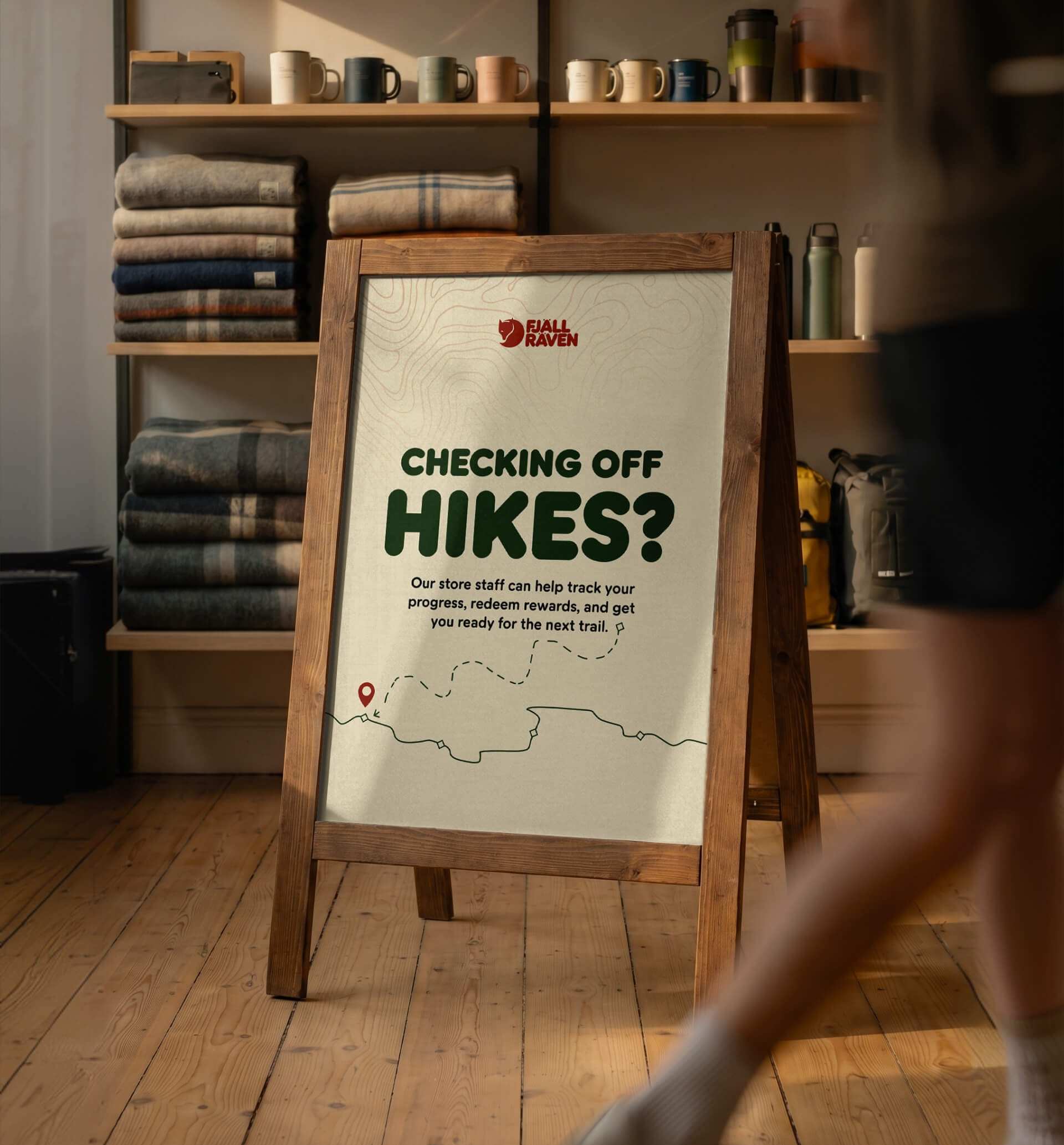 A-frame sign reading "Checking off hikes?" placed inside a cozy Scandinavian store with a blurred figure walking past.