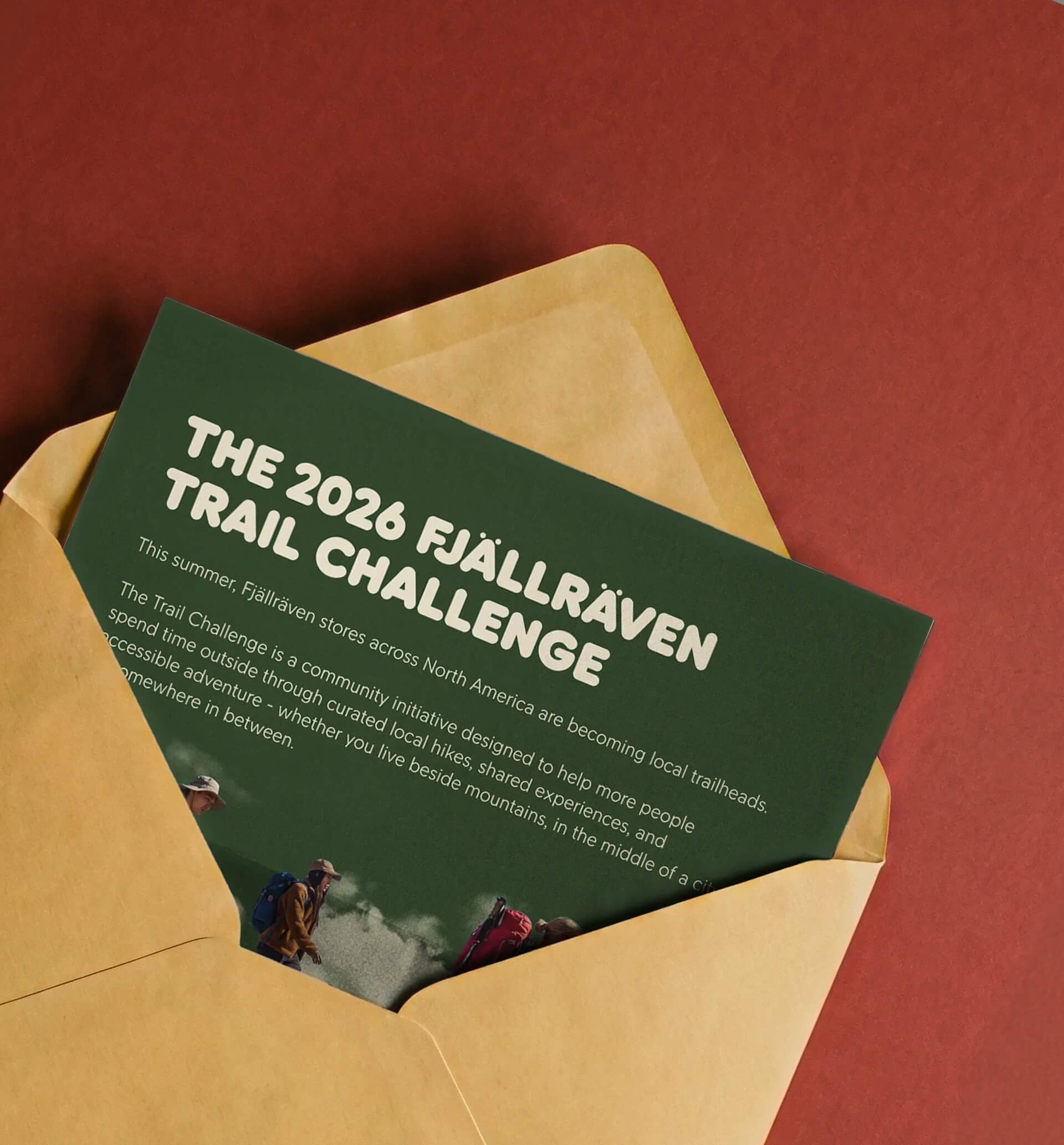 Forest green envelope holding a folded Trail Challenge invitation, on a warm sand surface.