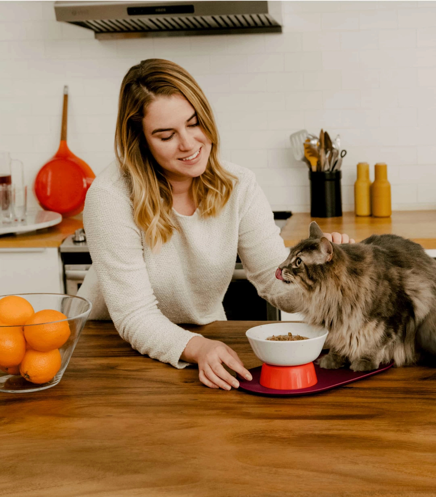 Smiling woman petting long-haired cat that is eating Cat Person wet food out of a Cat Person Mesa Bowl