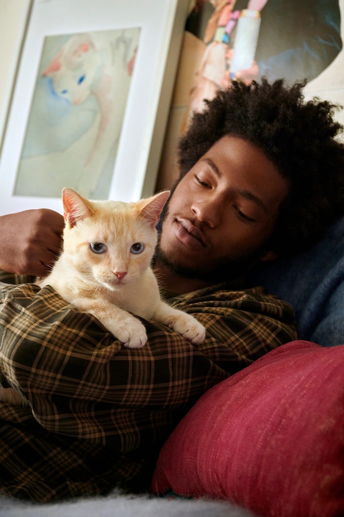 A man laying down on couch gazing at the cat he is holding in his arms. The cat peers out at the camera.