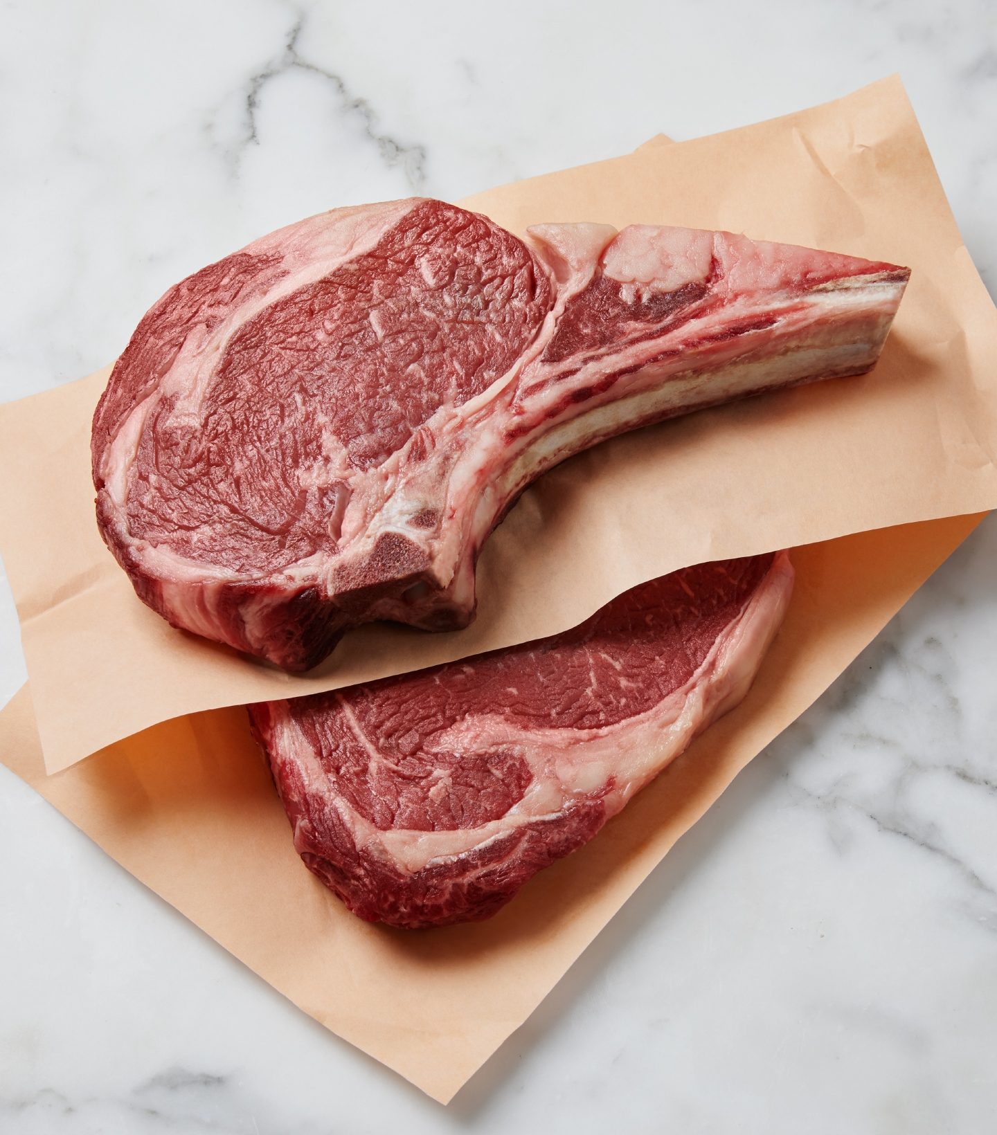 Two marbled raw beef ribeye steaks on a white marble countertop