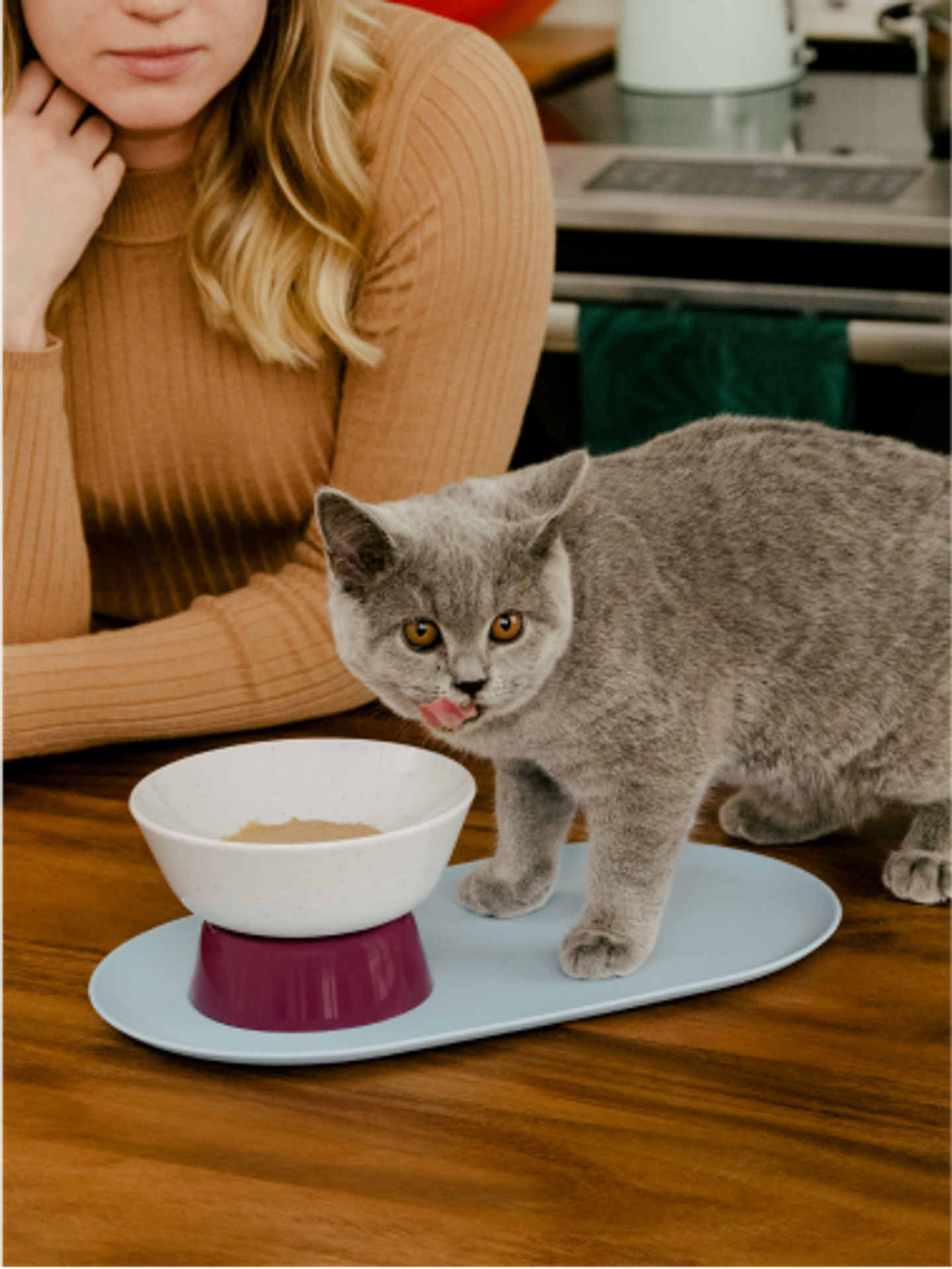 Maine coon kitten licking Cat Person Goodness Blends healthy cat treats out of a Cat Person Mesa Bowl on kitchen counter with woman looking from background
