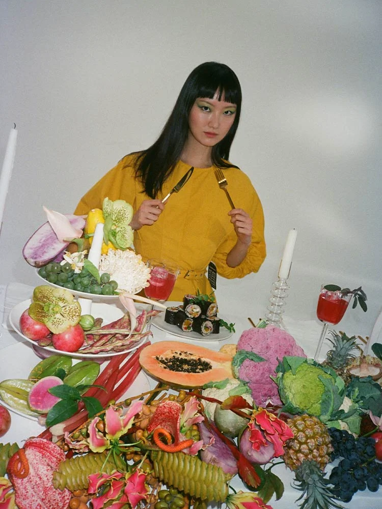 Behind the scenes campaign photo, female model in yellow surrounded by fruits and vegetables. Campaign Art direction by RoAndCo