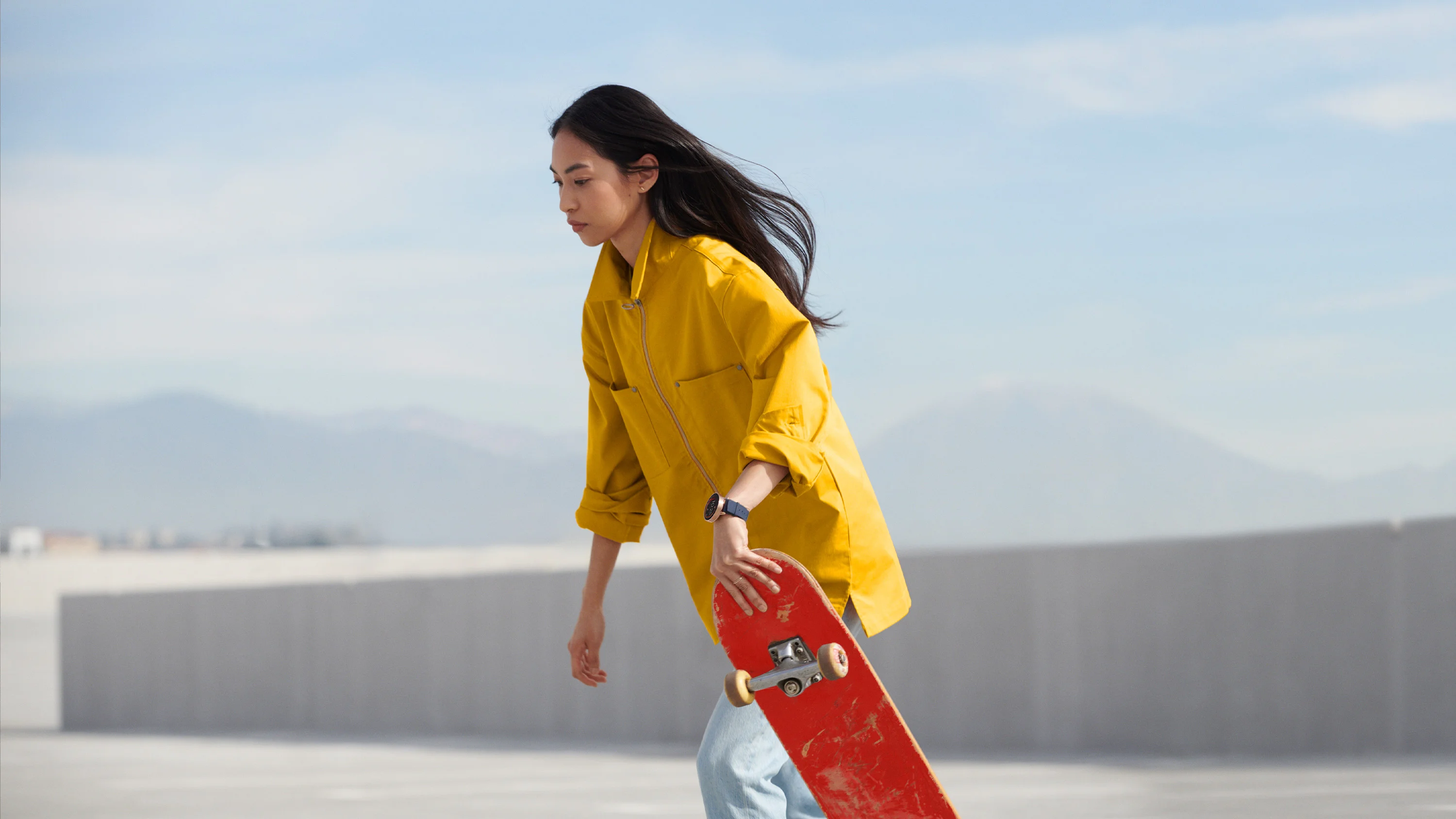 Woman in yellow jacket skateboarding, wearing black Google Wear OS watch, art direction by RoAndCo Studio