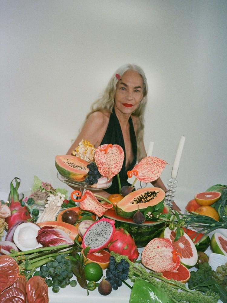 Behind the scenes campaign photo, female model in green, sitting at table surrounded by fruits and vegetables. Campaign Art direction by RoAndCo