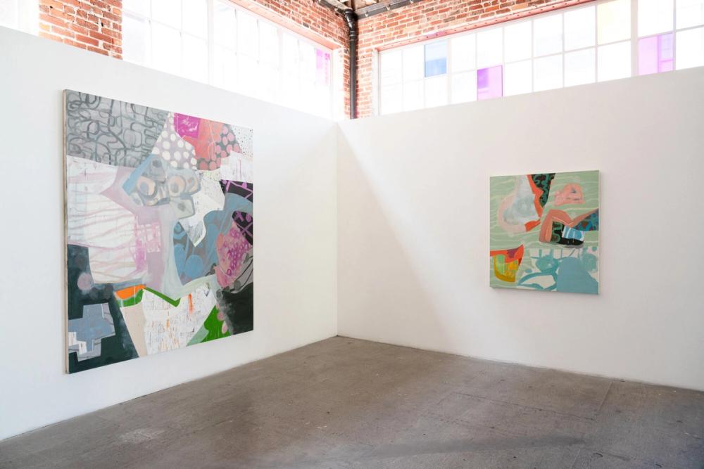 Two abstract paintings hang on white walls in an art gallery with brick and large windows in the background.
