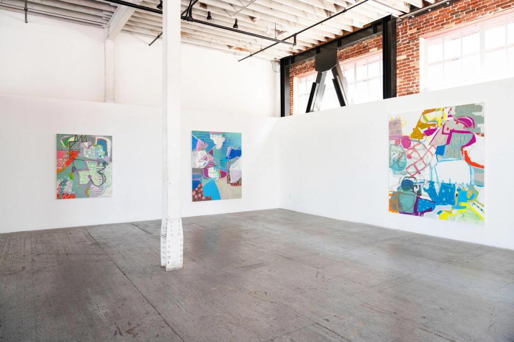 An art gallery with three colorful abstract paintings hanging on white walls in an industrial space with a brick wall and large windows.