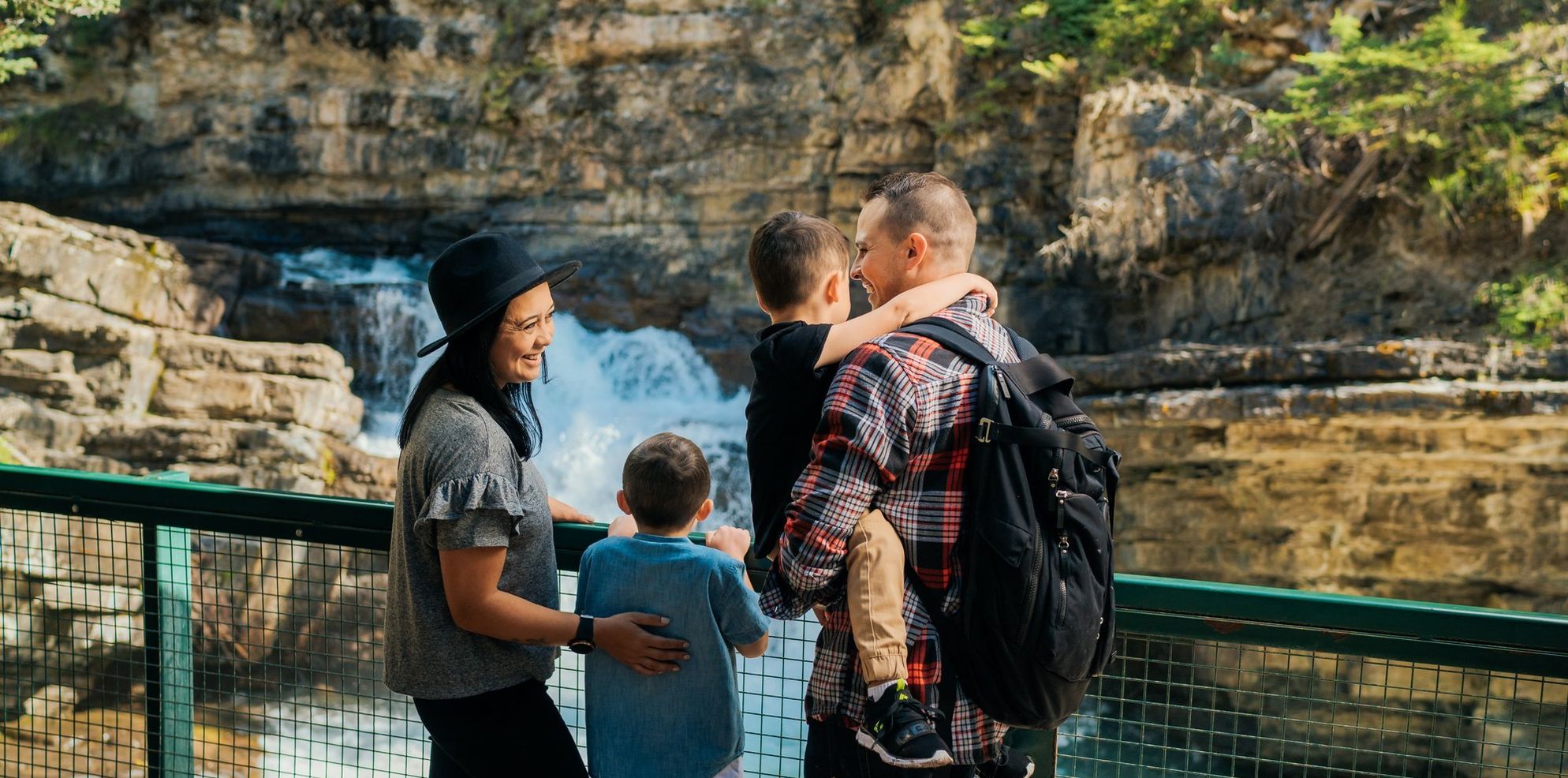A family of four stands at a railing overlooking a rushing waterfall as they laugh and embrace each other
