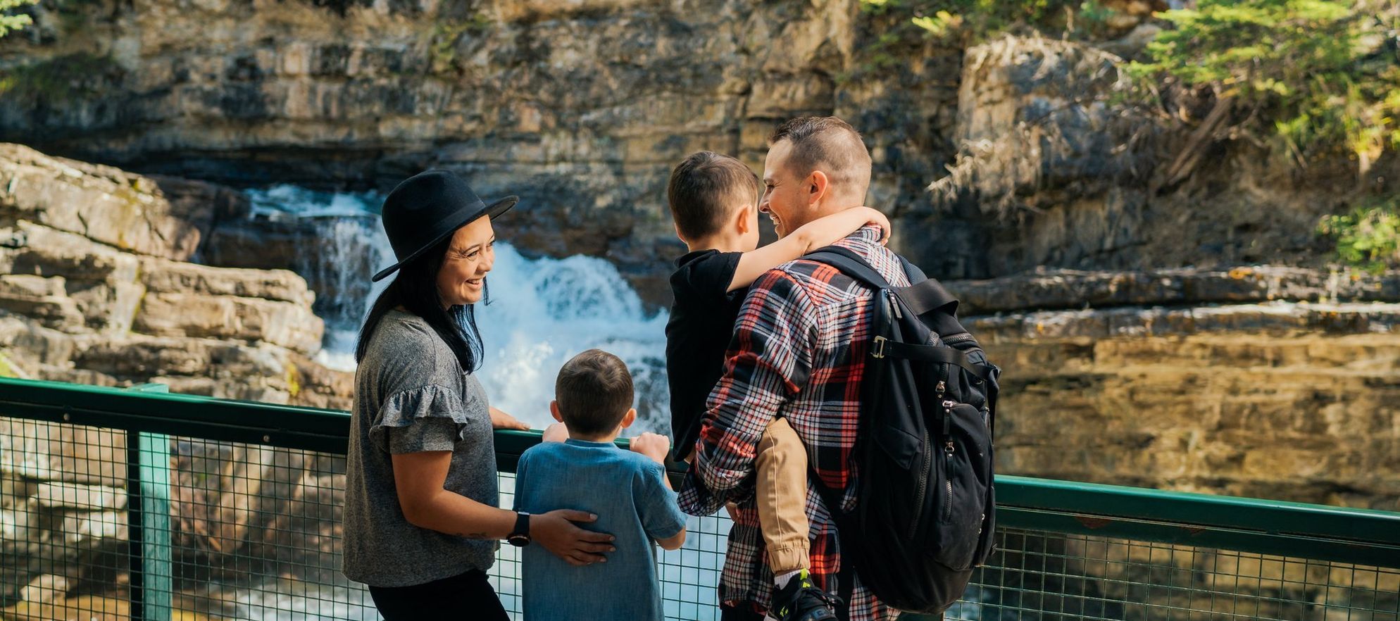 A family of four stands at a railing overlooking a rushing waterfall as they laugh and embrace each other