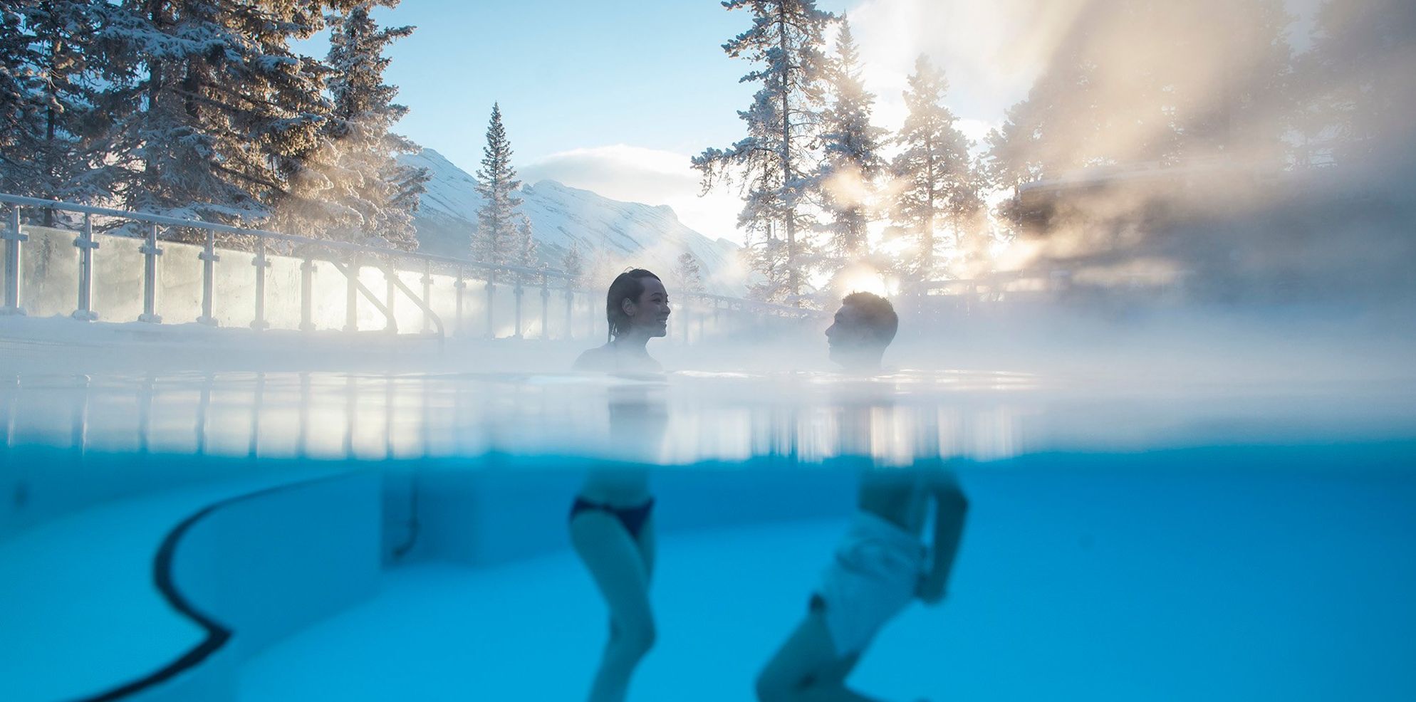 Two people in the Banff Upper Hot Springs in Banff National Park with sunlight pouring through the trees behind them.