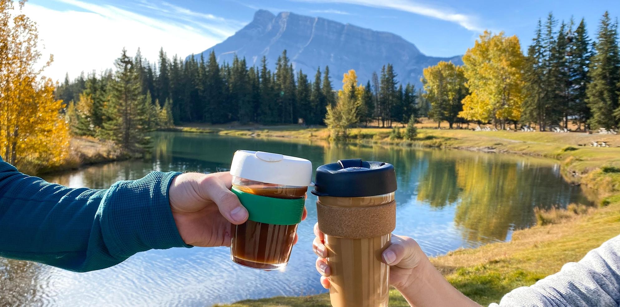 Two coffee cups "cheers-ing" at Cascade Ponds. Rundle Mountain and Fall colours in the background