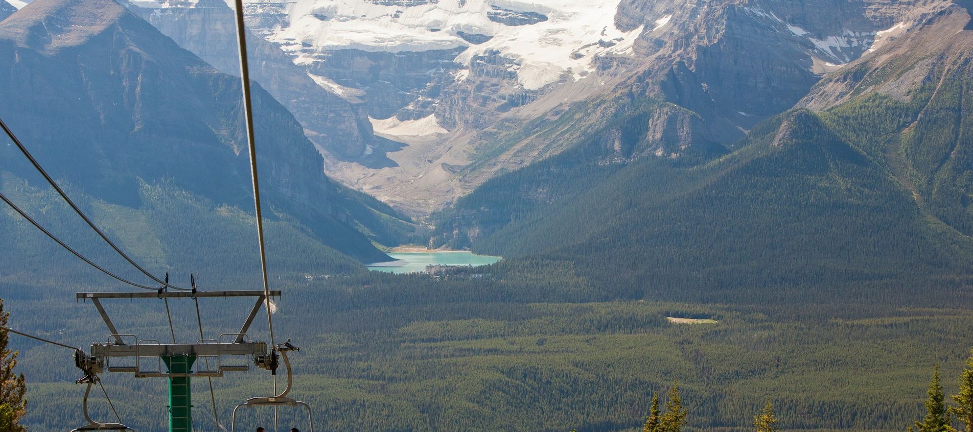 A couple travels down a mountain in an open chairlift while taking in the view of Lake Louise and Victoria Glacier in the distance
