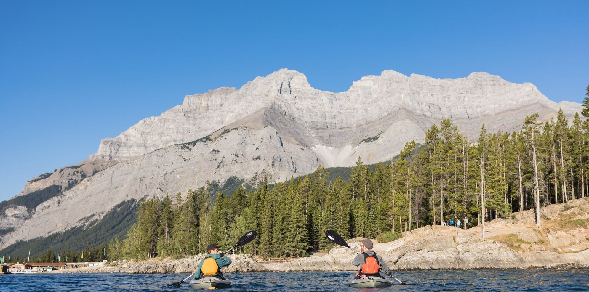 Two young males paddle in kayaks on a large blue lake with mountains and trees surrounding