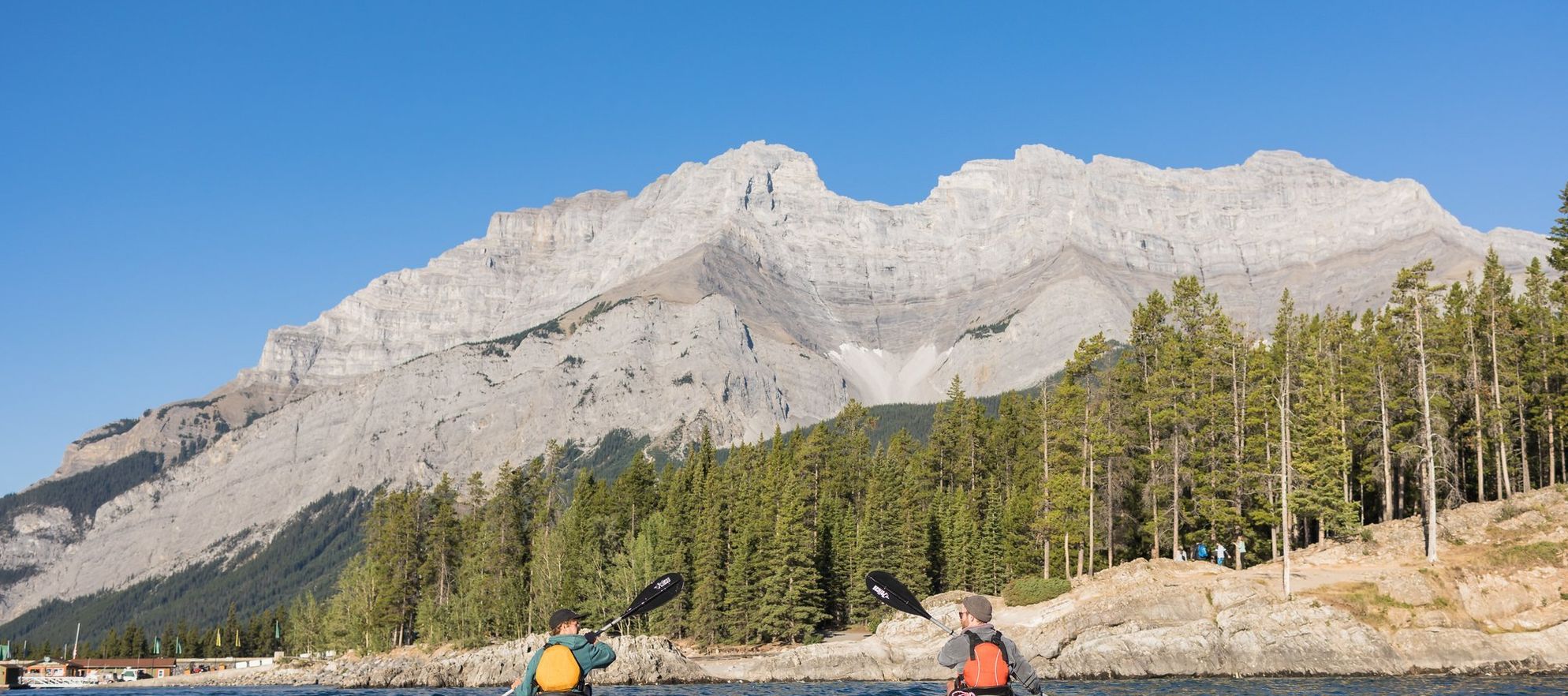 Two young males paddle in kayaks on a large blue lake with mountains and trees surrounding