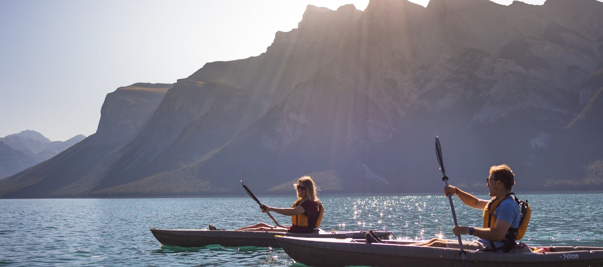 Two friends paddle in kayaks along a stunning turquoise lake with a clear blue sky and sunny sky