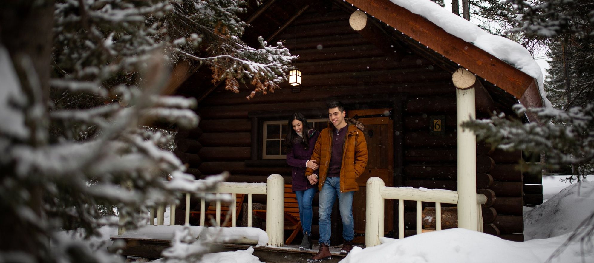 A couple in front of a cozy winter cabin surrounded by snowy trees in Banff National Park.