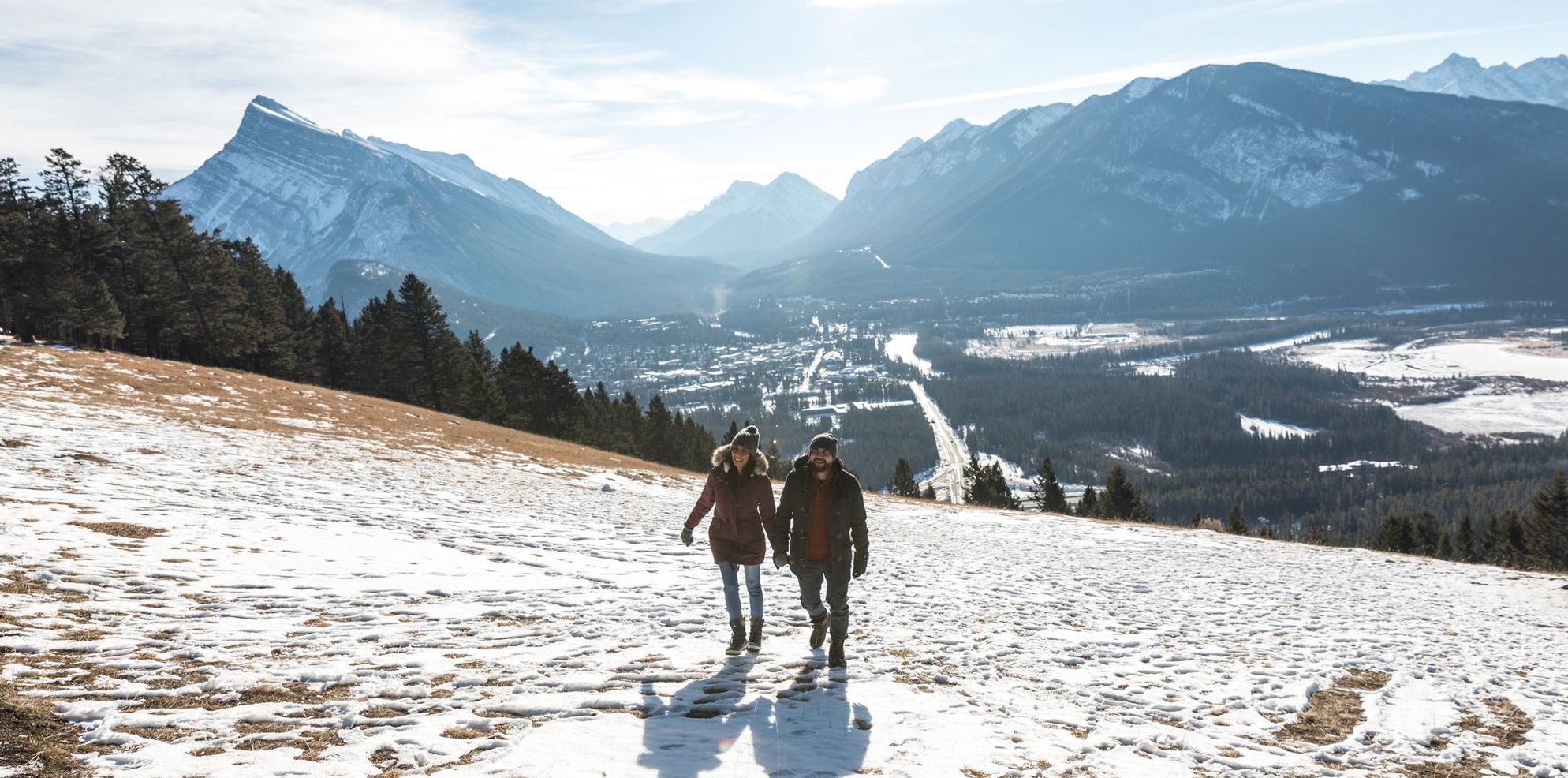 A couple hiking on a hill with grass peeking through the snow and an aerial view of the town and the mountains behind them