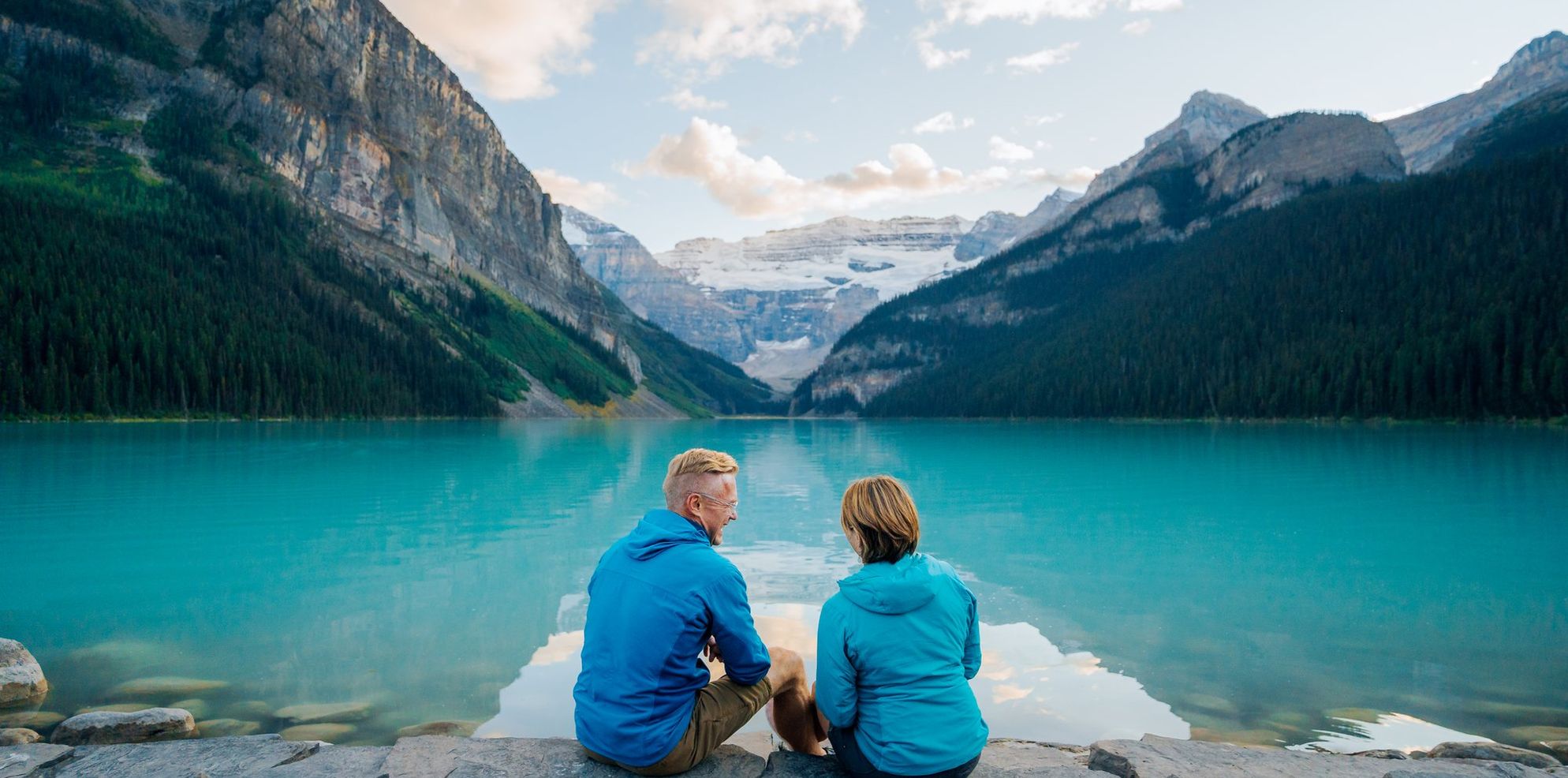 A mature couple sits on the rocks at the edge of a vast turquoise blue lake with a glacier and large mountains in view at the end of the lake