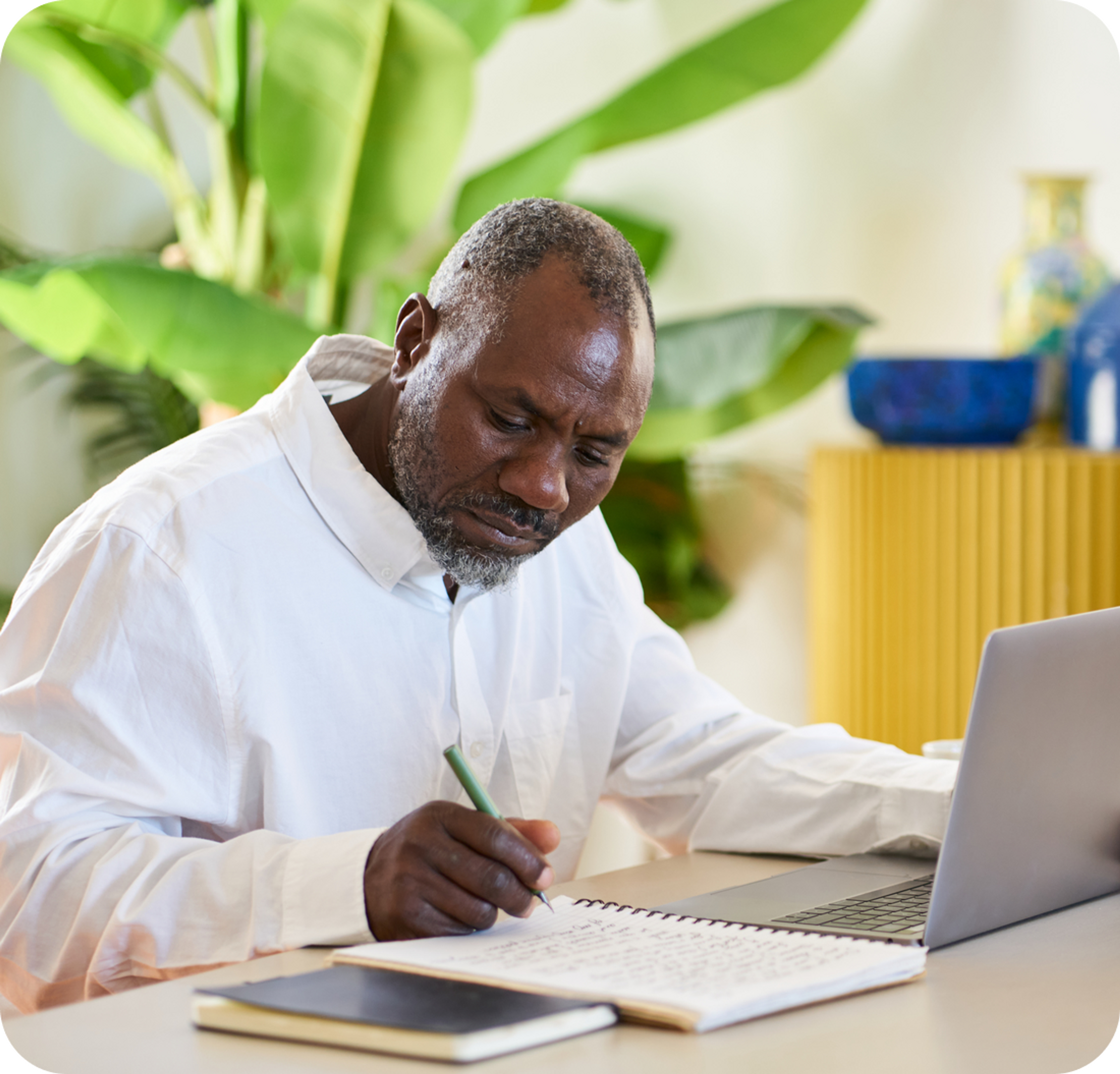 A photo of a man sat at a desk in front of a laptop and writing on a notepad