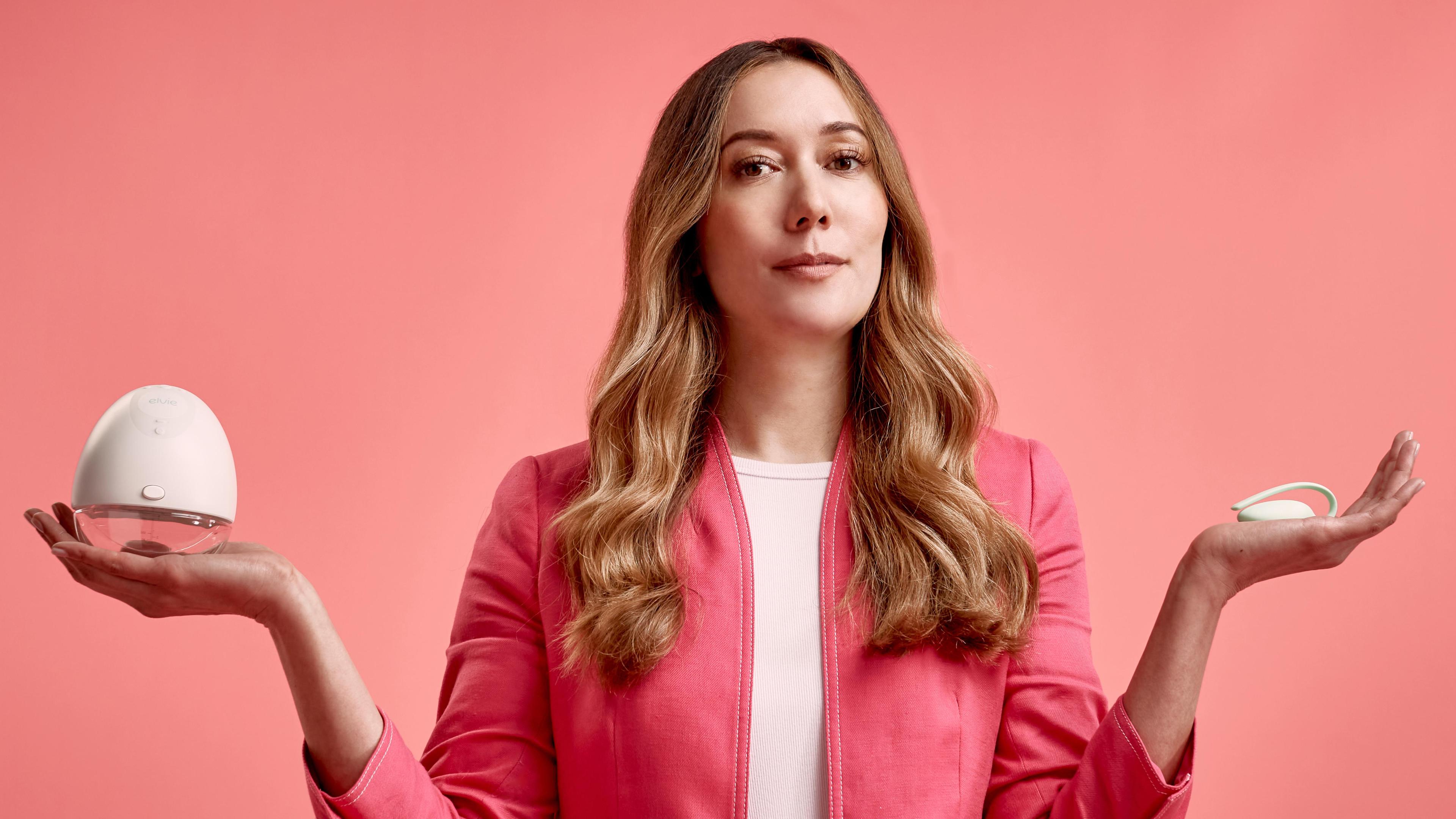 A woman in a pink blazer over a lighter pink top stands against a coral pink background, holding a white egg-shaped device on her left palm and a small circular device on her right palm. Her expression is neutral.