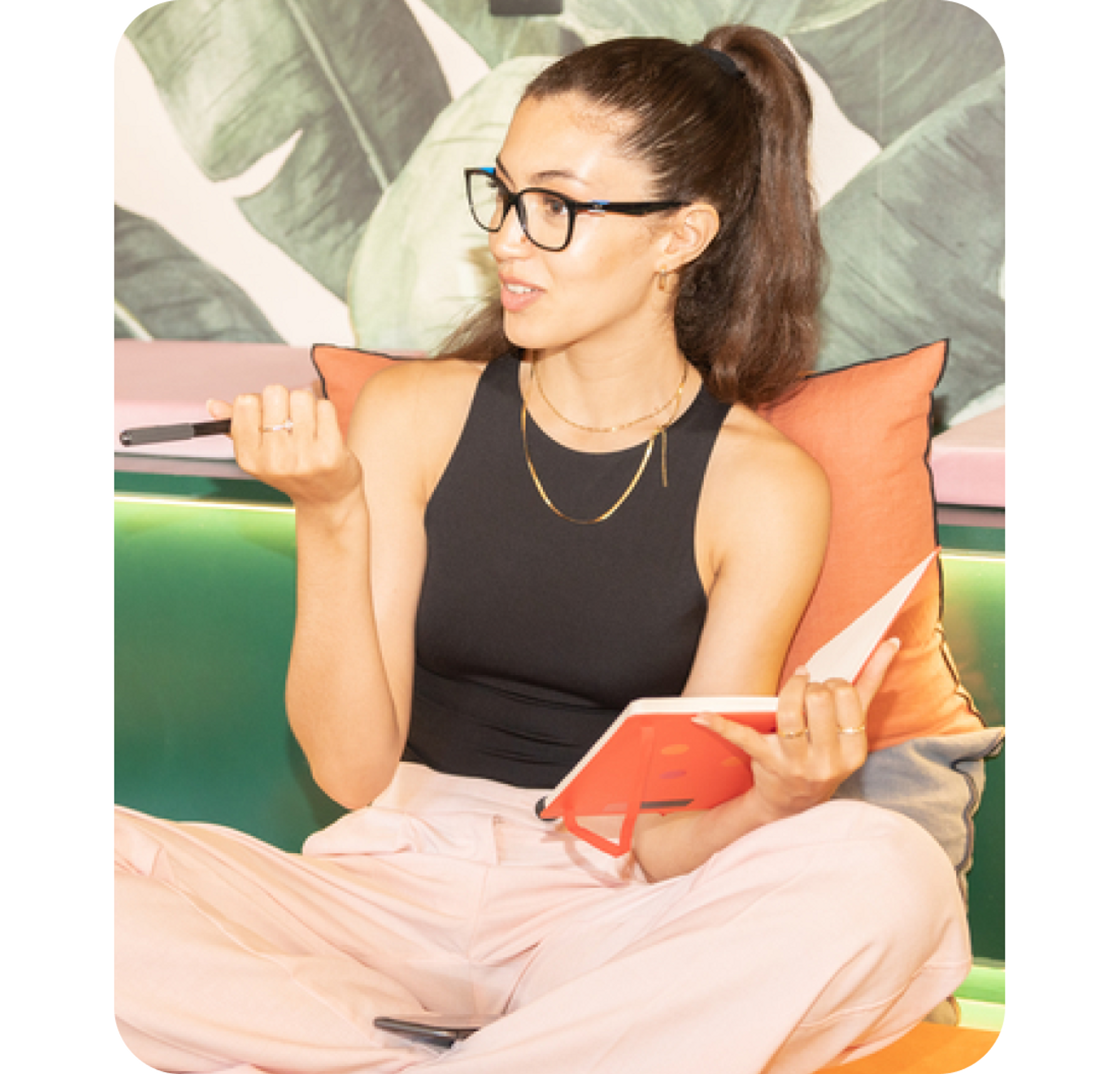 A photo of a smiling woman sat down on a sofa and holding a notebook