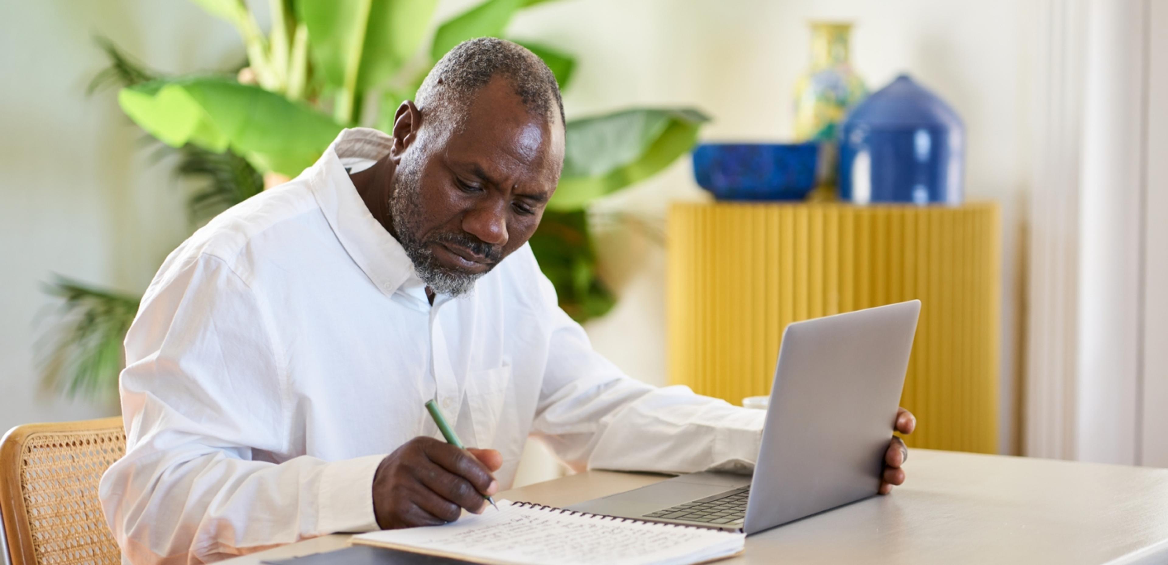 An older Black man in a white shirt is focused on writing in a notebook while sitting at a white table with a laptop open in front of him. The room is bright with natural light, and the decor includes a potted plant and a blue vase on a yellow cabinet, suggesting a comfortable home environment.