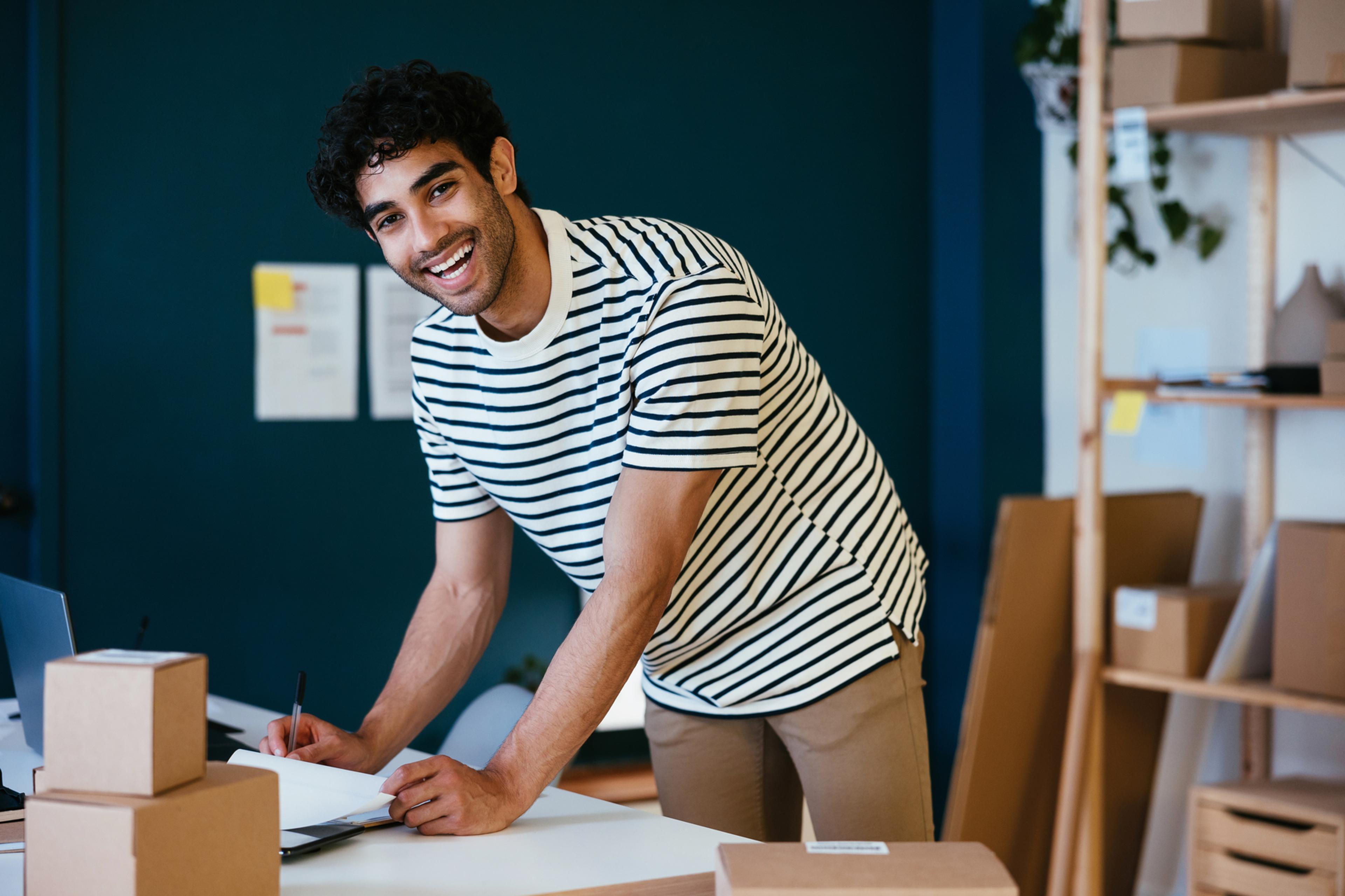 A photo of a man smiling while stood up and bent over a desk with paperwork