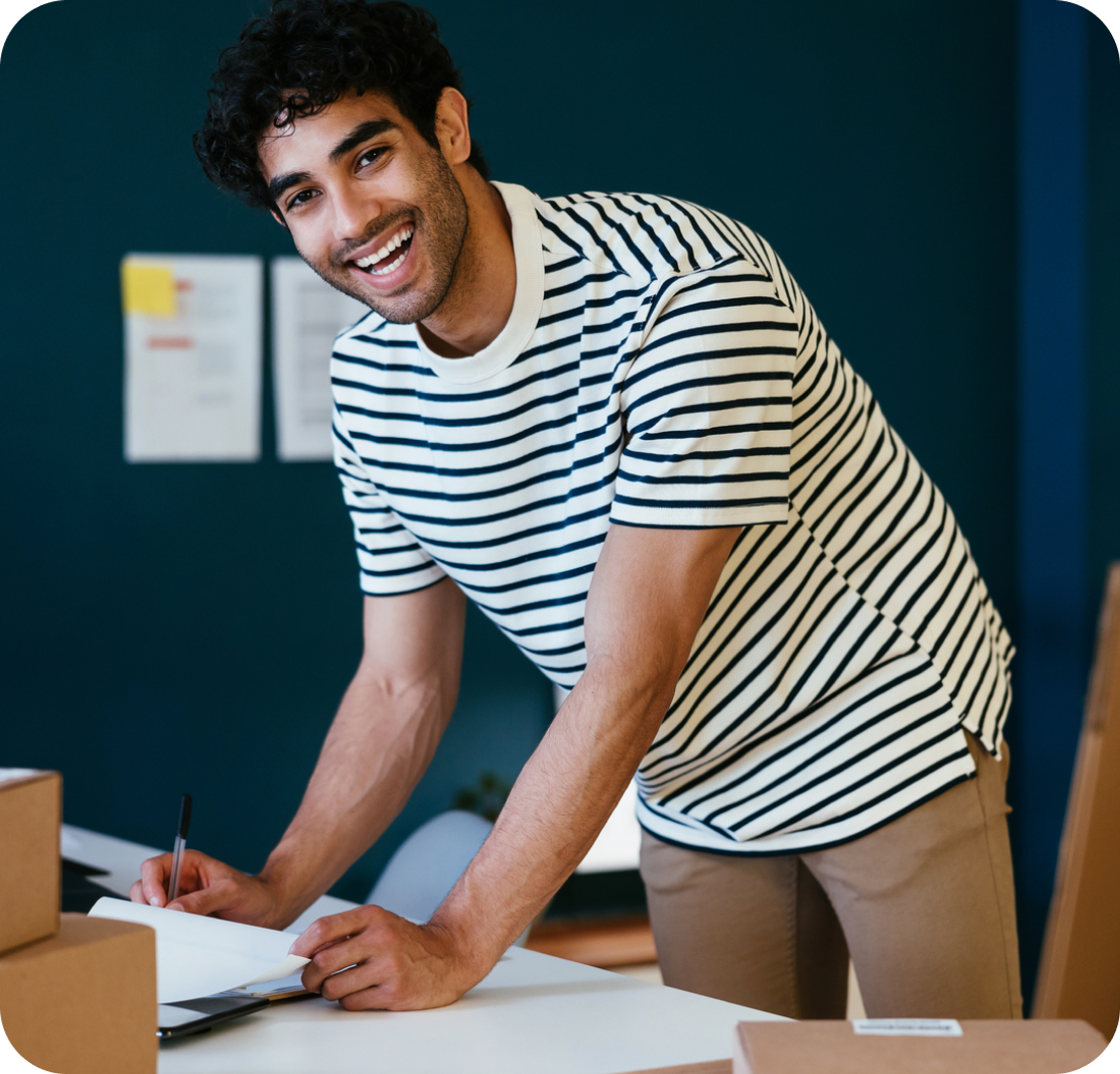 A photo of a man smiling and bent over a desk while holding a pen