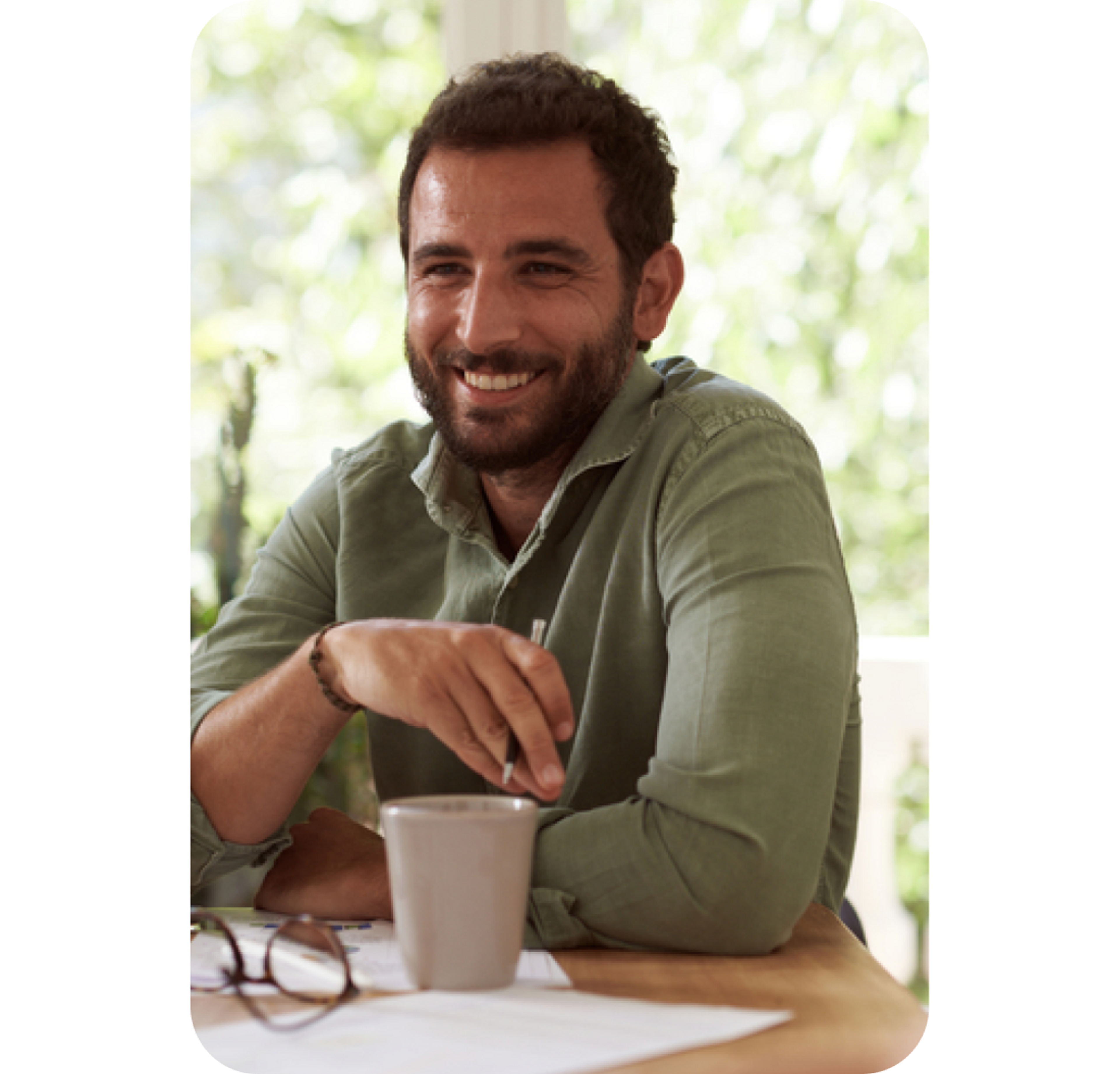 A photo of a man smiling while sat down and holding a pen