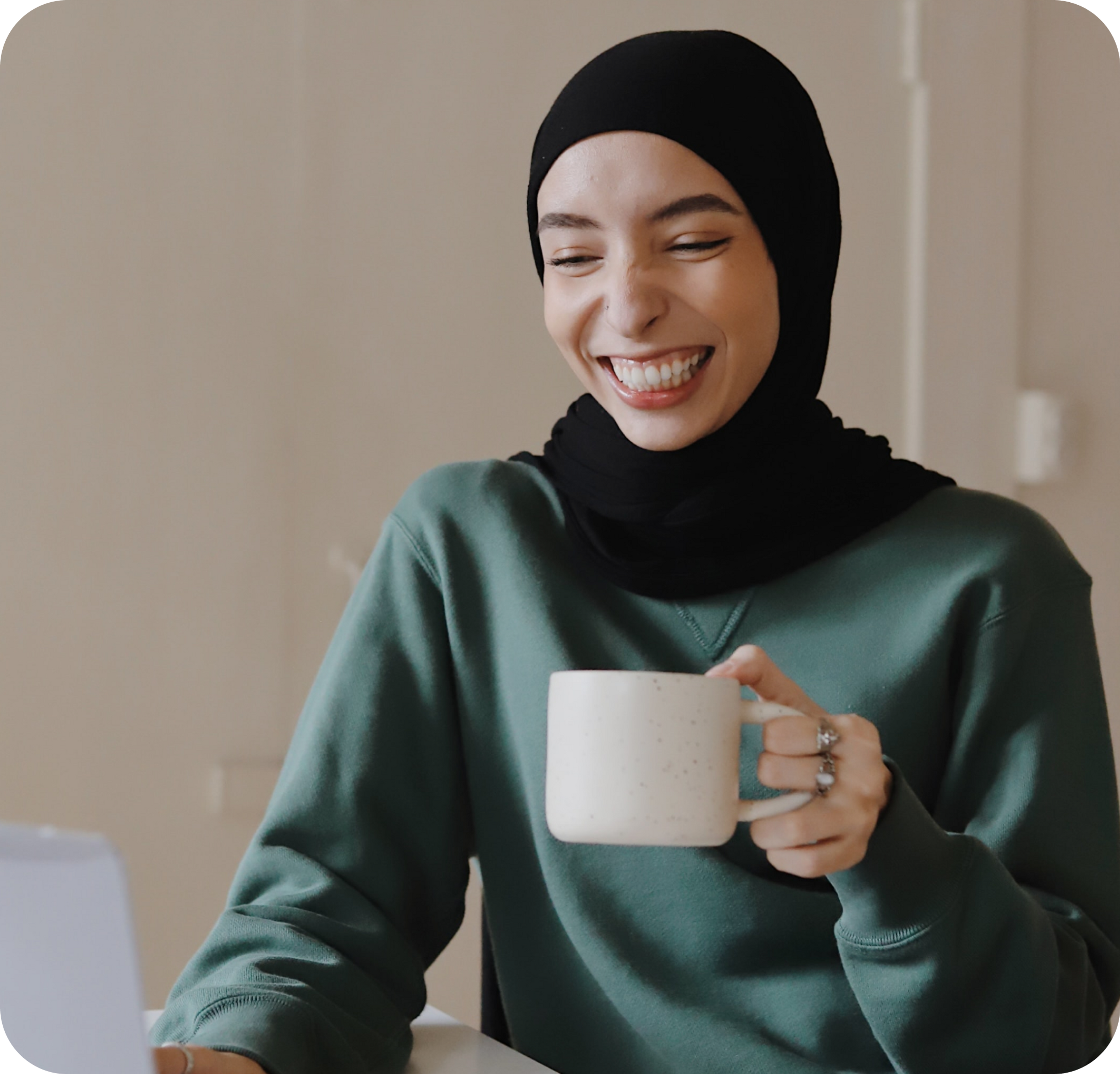 A photo of a woman smiling while holding a mug and looking at a laptop