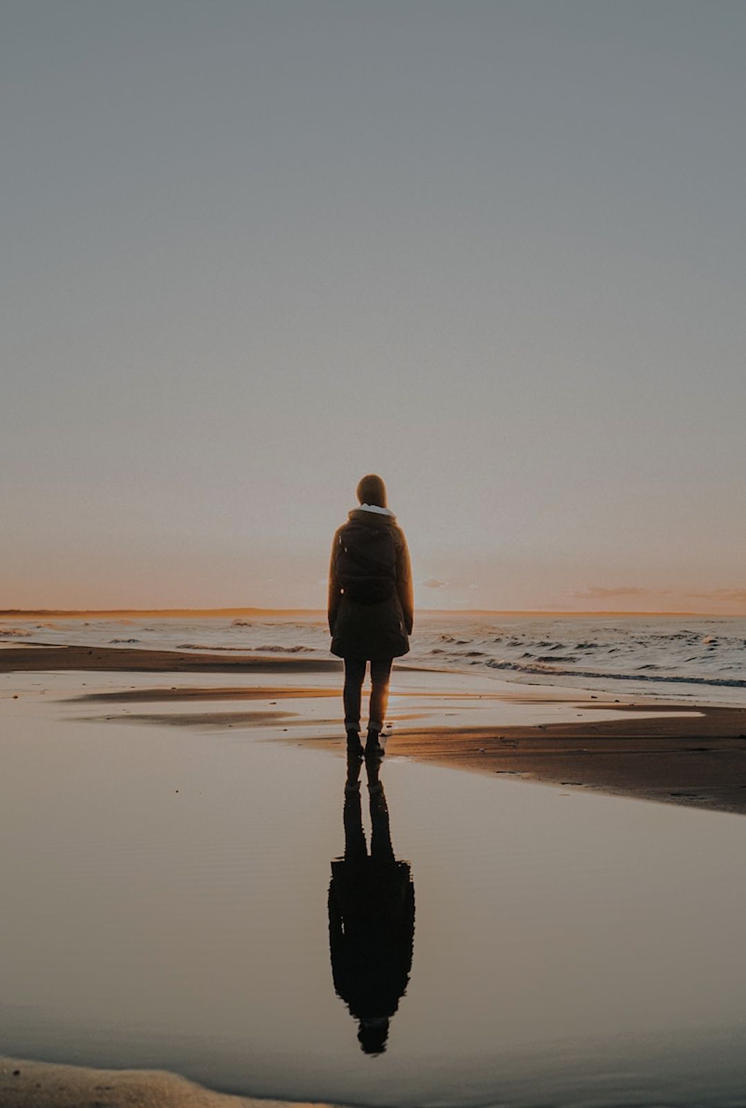 Person walking alone along a reflective beach at sunset, with their silhouette mirrored in the shallow water