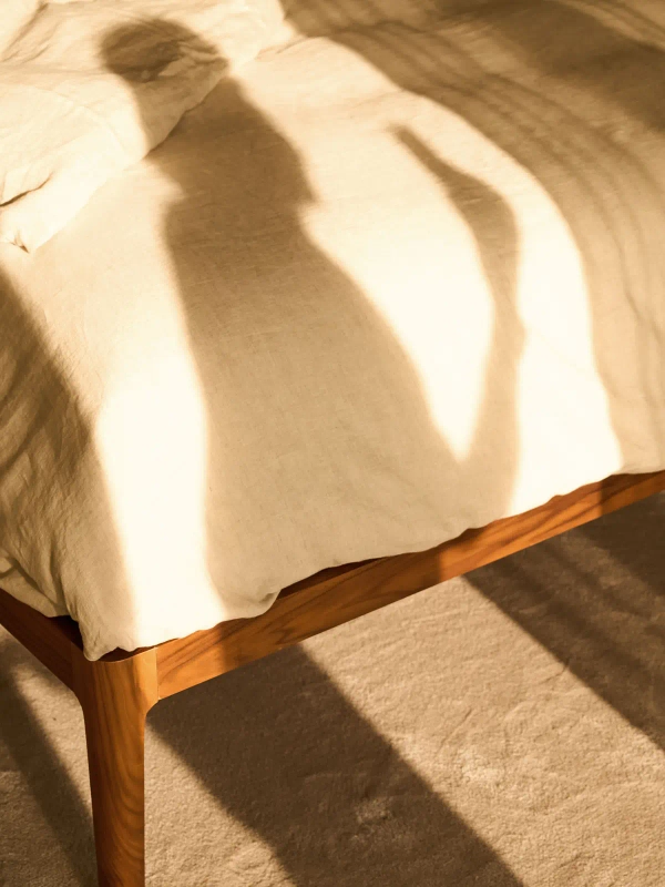 Abstract shadows fall across a sunlit linen bed and wooden frame.