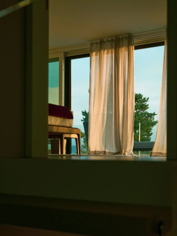 A bright room with sheer curtains and a serene outdoor view of trees and sky is seen through a dark, framed doorway or opening.