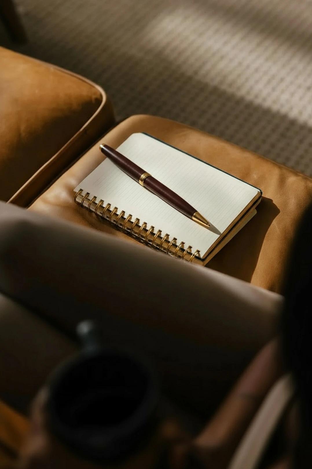 A closed spiral-bound notebook with a dark pen resting on its cover lies on a sunlit brown leather cushion, creating a cozy scene.