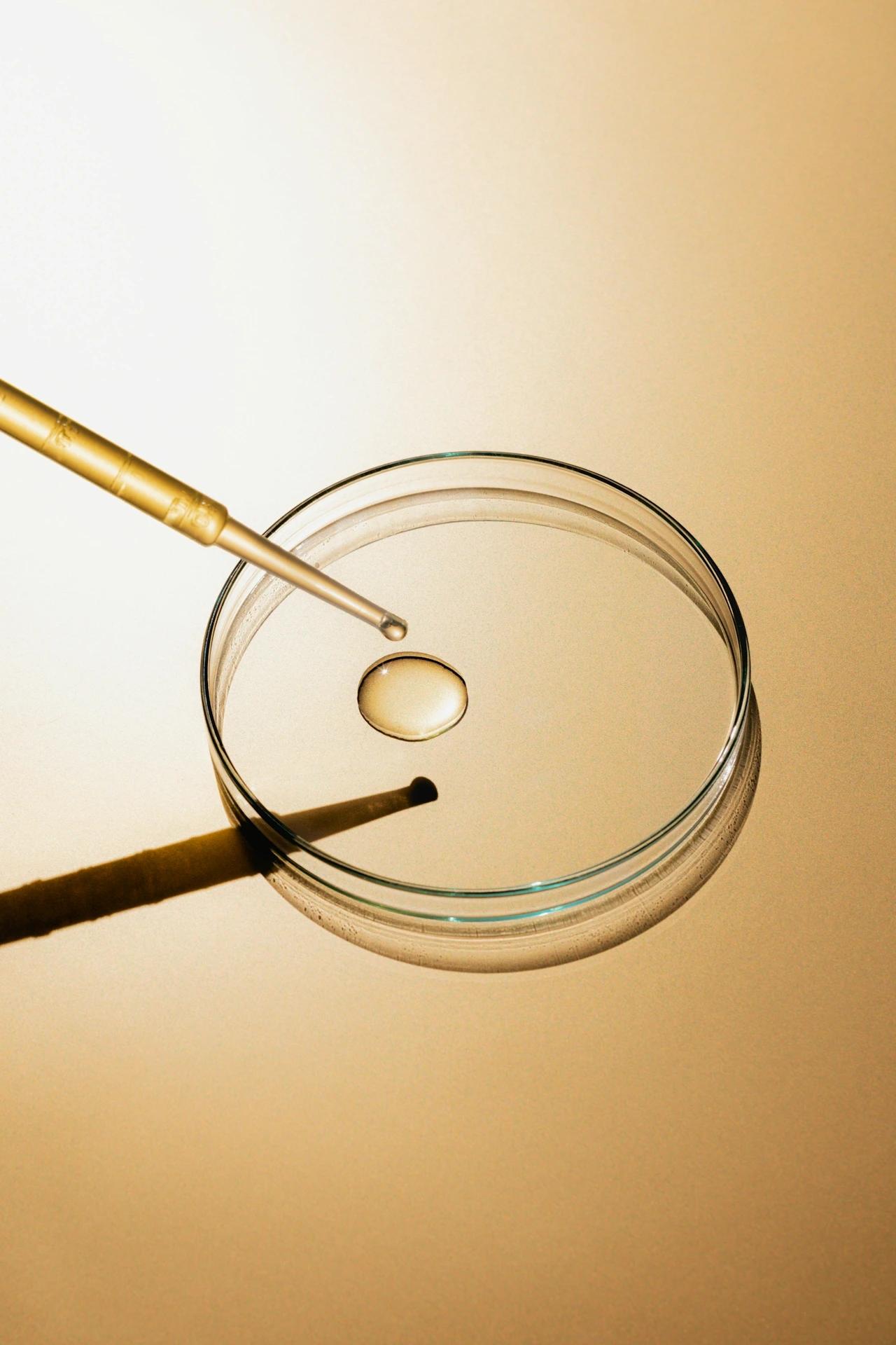 A golden dropper dispenses a clear liquid droplet into a glass petri dish, set against a warm, reflective golden background.